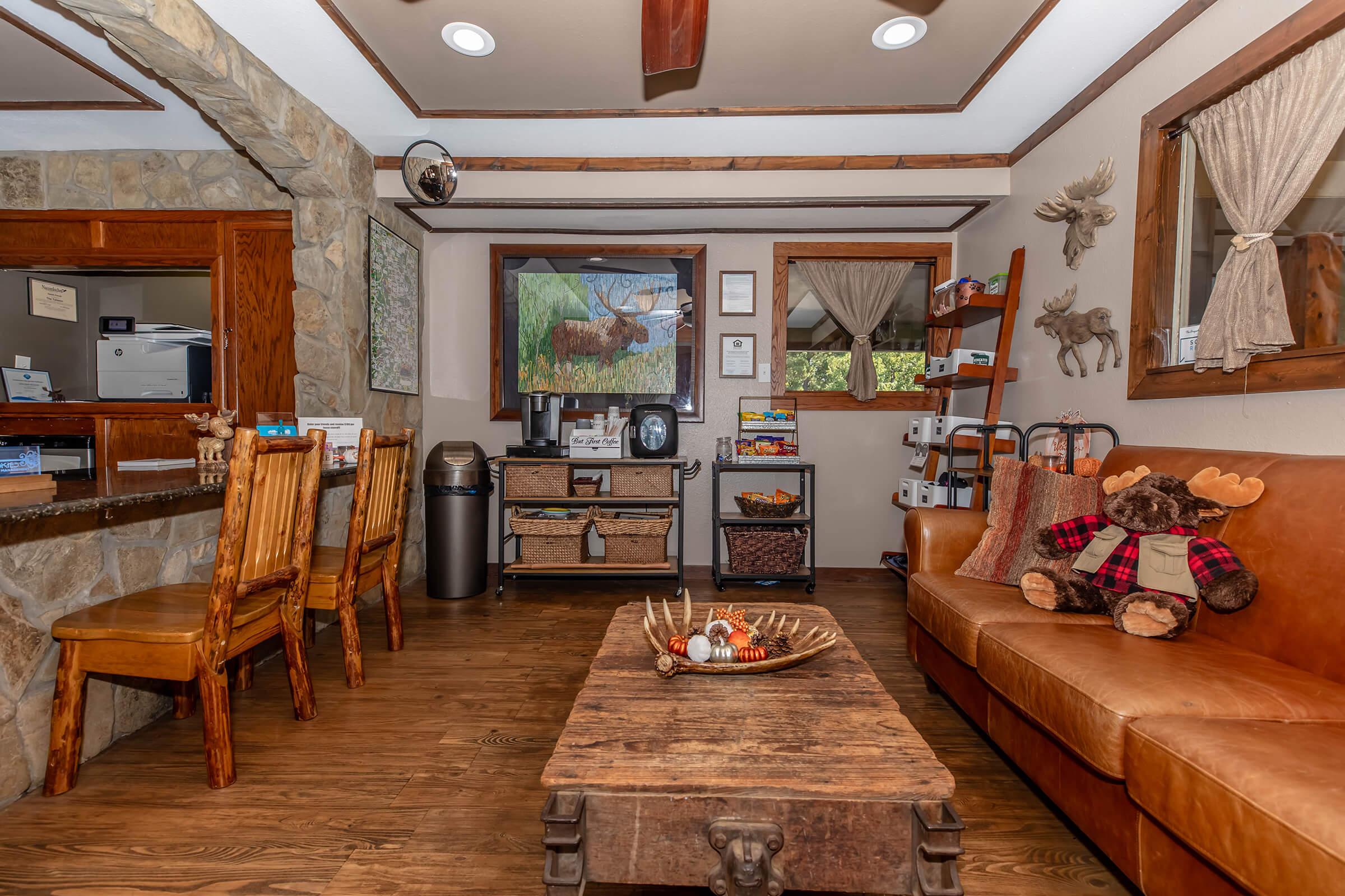 Cozy interior of a rustic waiting area featuring a brown leather sofa, wooden chairs, and a coffee table with decorative items. A stone wall is visible, along with a reception desk, storage cart, and wildlife-themed decor. Large windows offer a view of nature, creating a warm and inviting atmosphere.