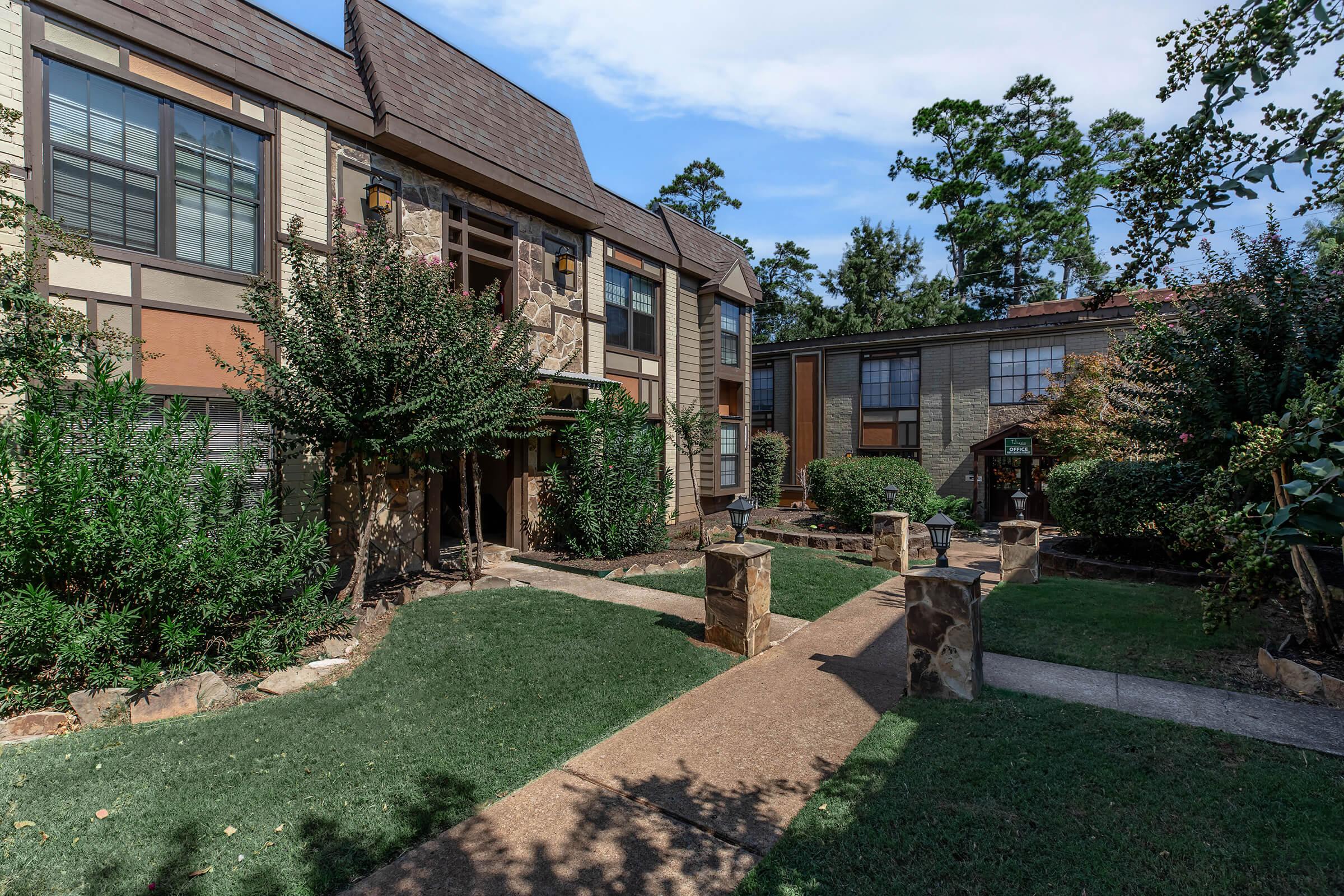 A well-maintained apartment community surrounded by lush greenery, featuring a stone walkway leading through landscaped gardens. The buildings exhibit a combination of brick and wooden siding with large windows, and decorative outdoor lamps add charm to the setting. The sky is clear with a hint of clouds.