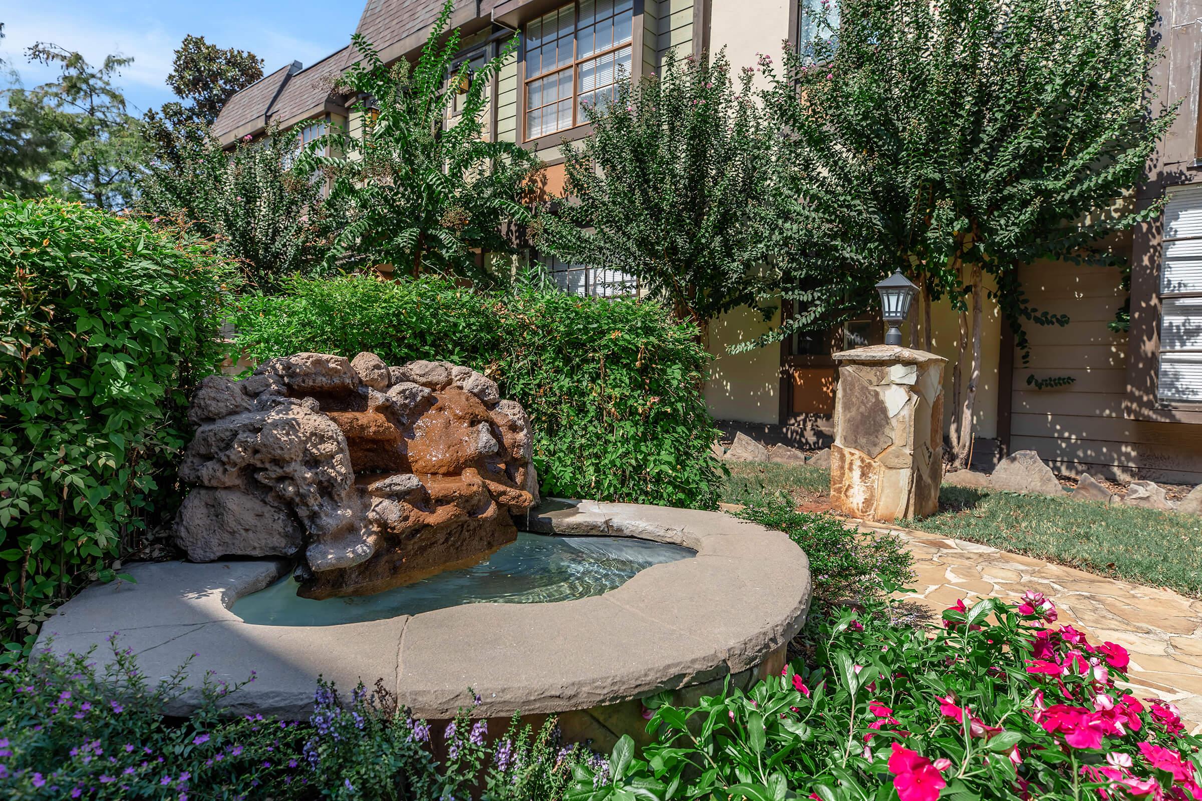 A tranquil garden scene featuring a small stone fountain with gently flowing water, surrounded by lush greenery and colorful flower beds. The fountain is nestled against a backdrop of a charming building with large windows and well-maintained landscaping, creating a peaceful outdoor atmosphere.