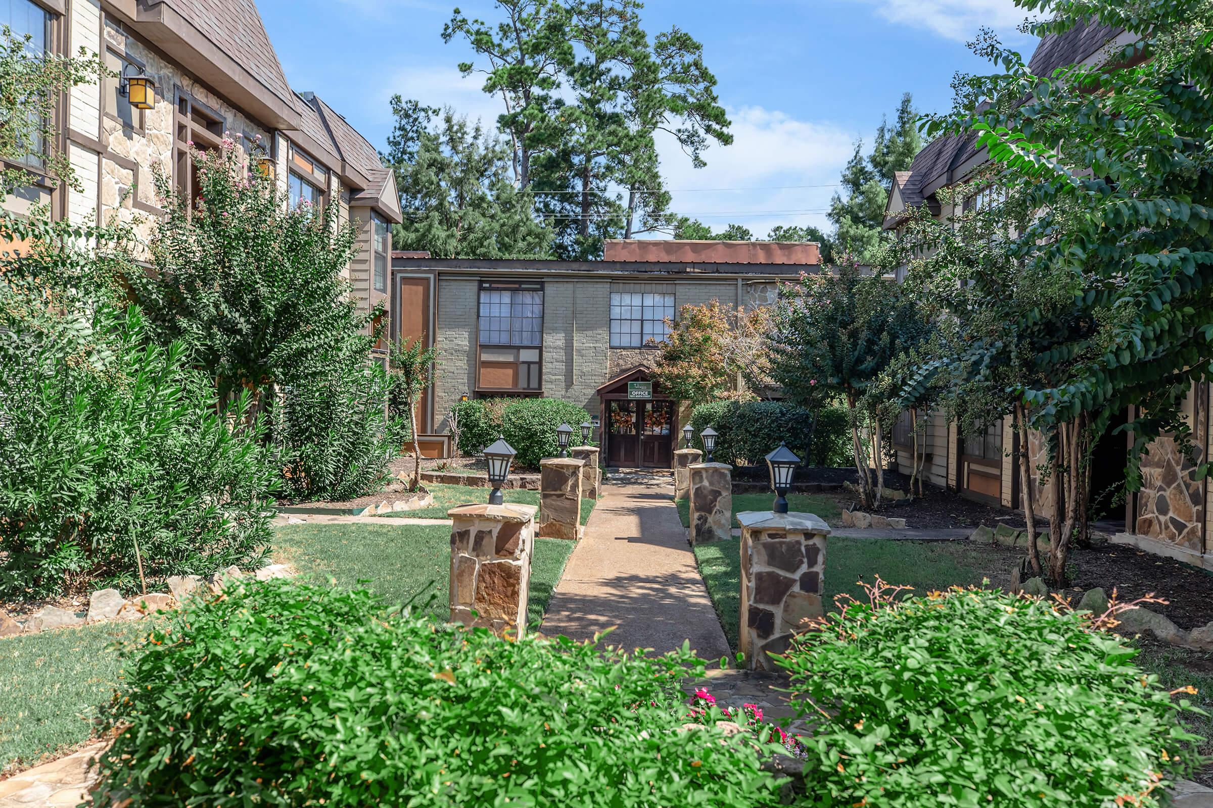 A landscaped courtyard featuring stone pathways, lush greenery, and decorative lanterns, framed by multi-story buildings. The scene is set under a clear blue sky with a few scattered clouds, creating a serene and inviting atmosphere.