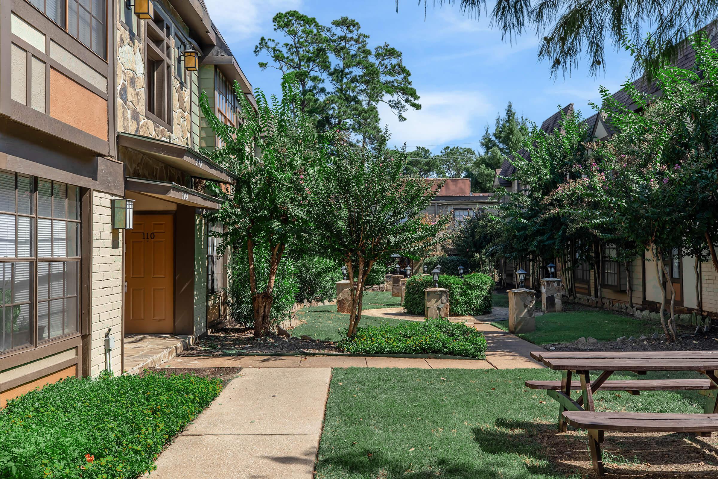 A landscaped courtyard featuring green lawns, shrubs, and trees. There are stone pathways leading to brown doors of two-story buildings. Picnic tables sit on the lawn, surrounded by lush greenery under a clear blue sky. The setting suggests a peaceful residential area or community space.