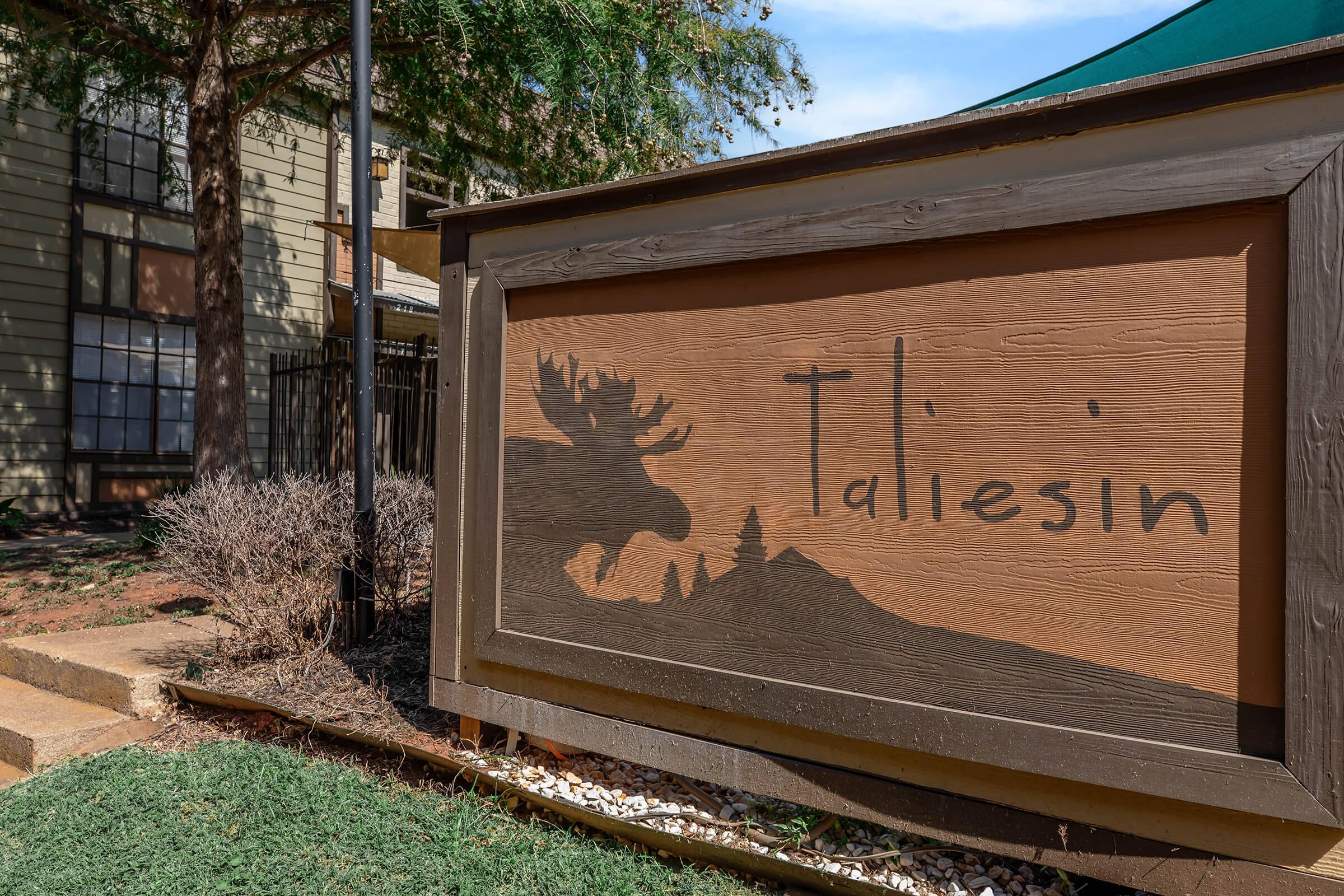 A rustic wooden sign for "Taliesin," featuring a silhouette of a moose with a forested mountain backdrop. The sign is mounted outside a building, surrounded by grass and small plants. Trees are visible in the background, indicating a natural setting.