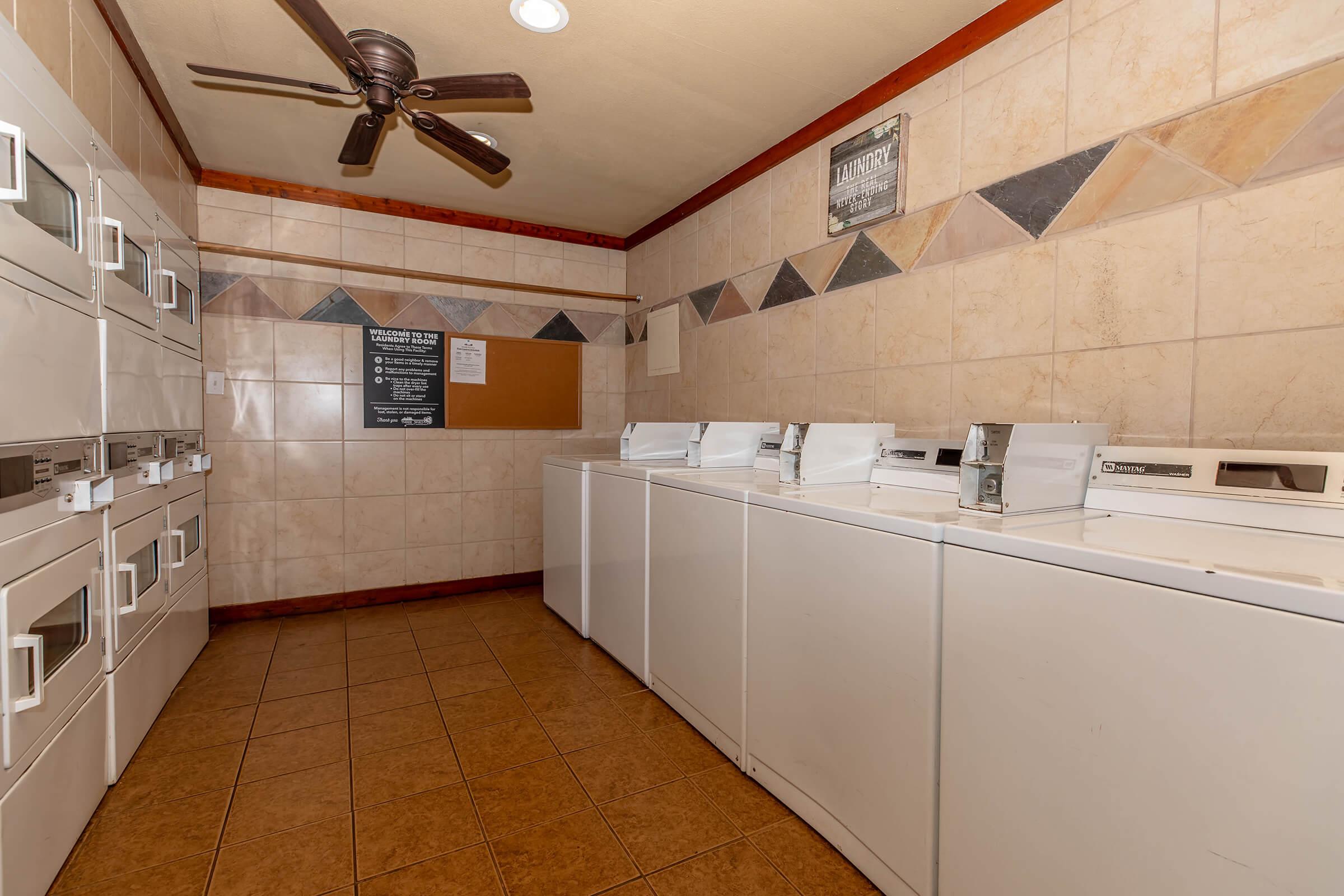 A laundry room featuring several white washing machines and dryers arranged in a row. The room has beige tiled walls with decorative stone accents and a ceiling fan. A bulletin board is mounted on the wall, and a sign with laundry instructions is visible. The floor is tiled with brown square tiles.