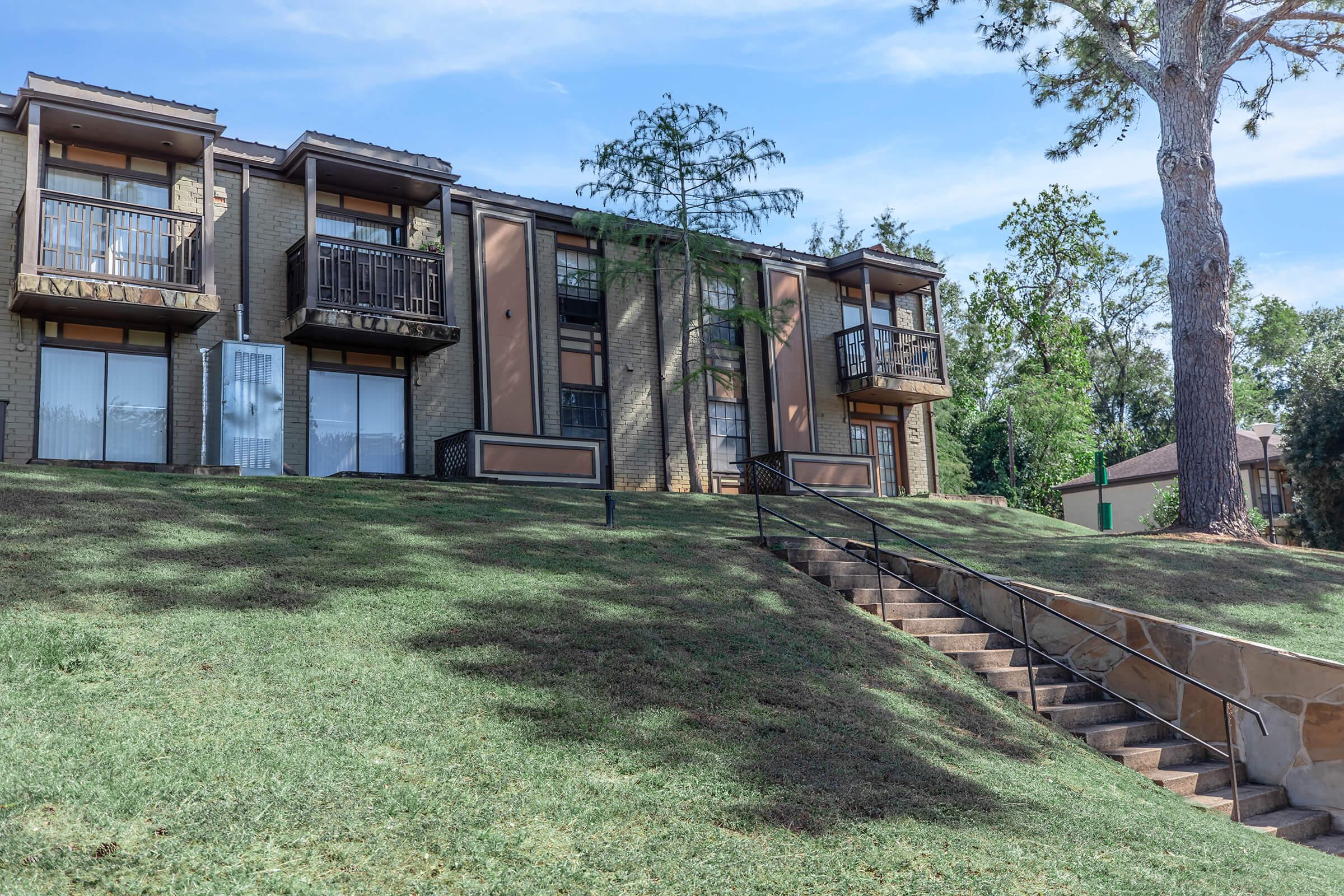 A multi-story residential building with balconies on each floor, situated on a grassy slope. Steps lead up from the lower ground level to the building entrance. Surrounding trees and shrubbery add a natural element to the scene, under a clear blue sky.