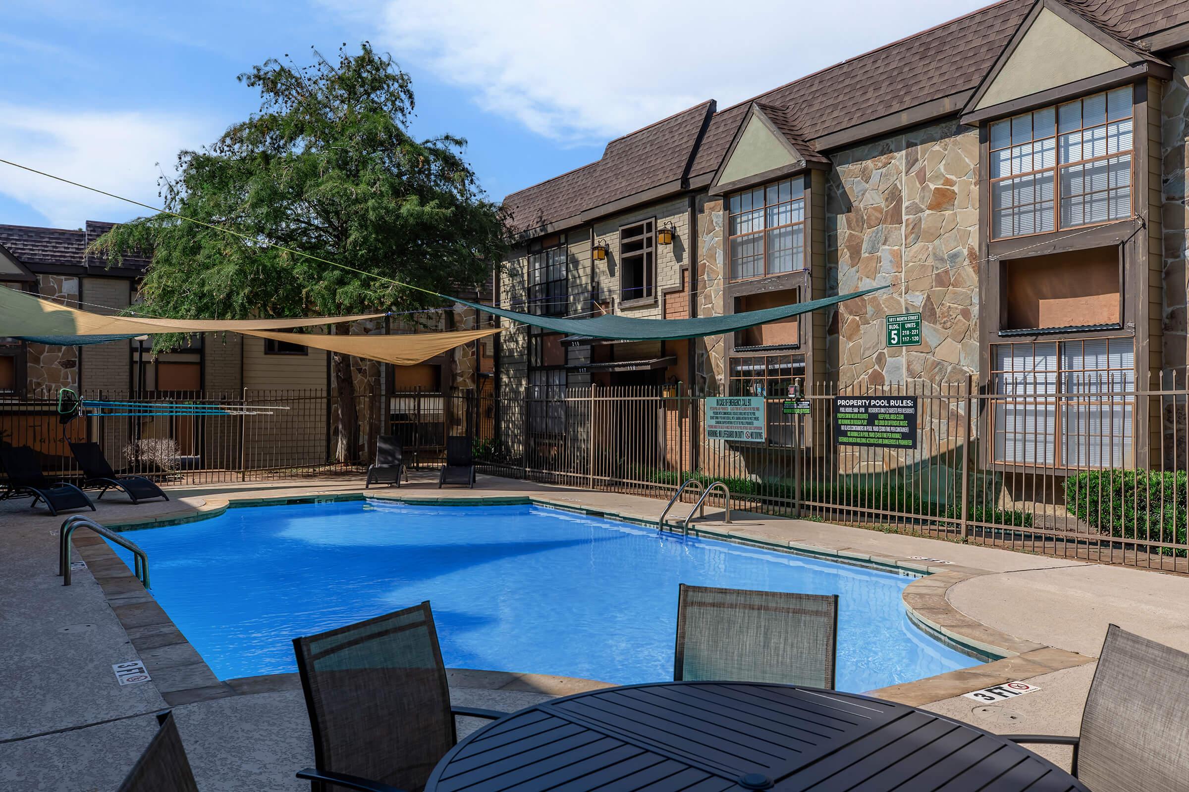 A clear blue swimming pool surrounded by lounge chairs and shaded by fabric canopies. The pool area is enclosed by a fence, with a few trees and a well-maintained apartment building in the background, featuring a stone facade. A sunny sky adds to the inviting atmosphere.