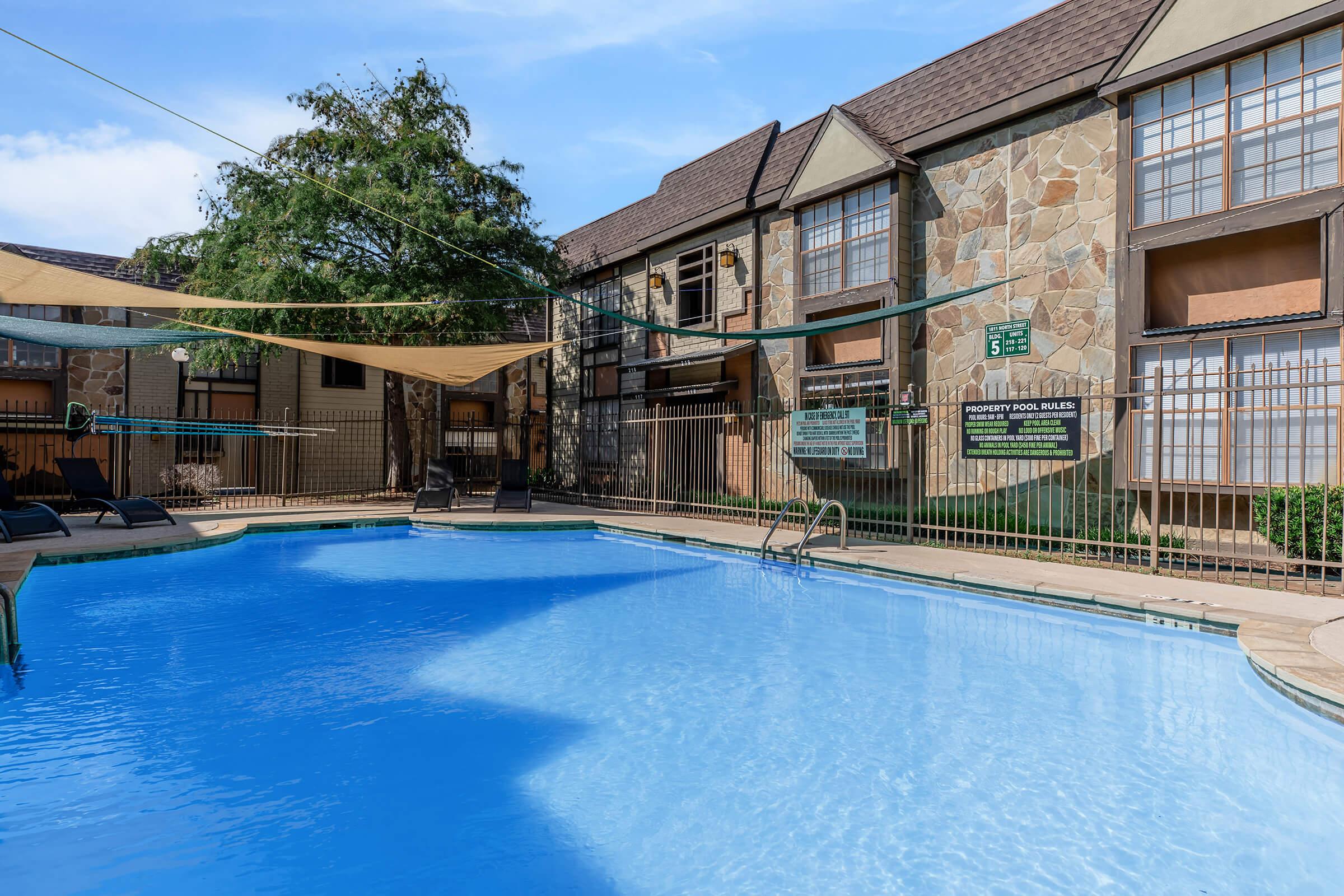 A clear blue swimming pool surrounded by a stone and wood-finished apartment building. There are lounge chairs around the pool, and shade sails overhead. A fence encloses the pool area, with signs displayed on the wall. Green trees are visible in the background under a bright sky.