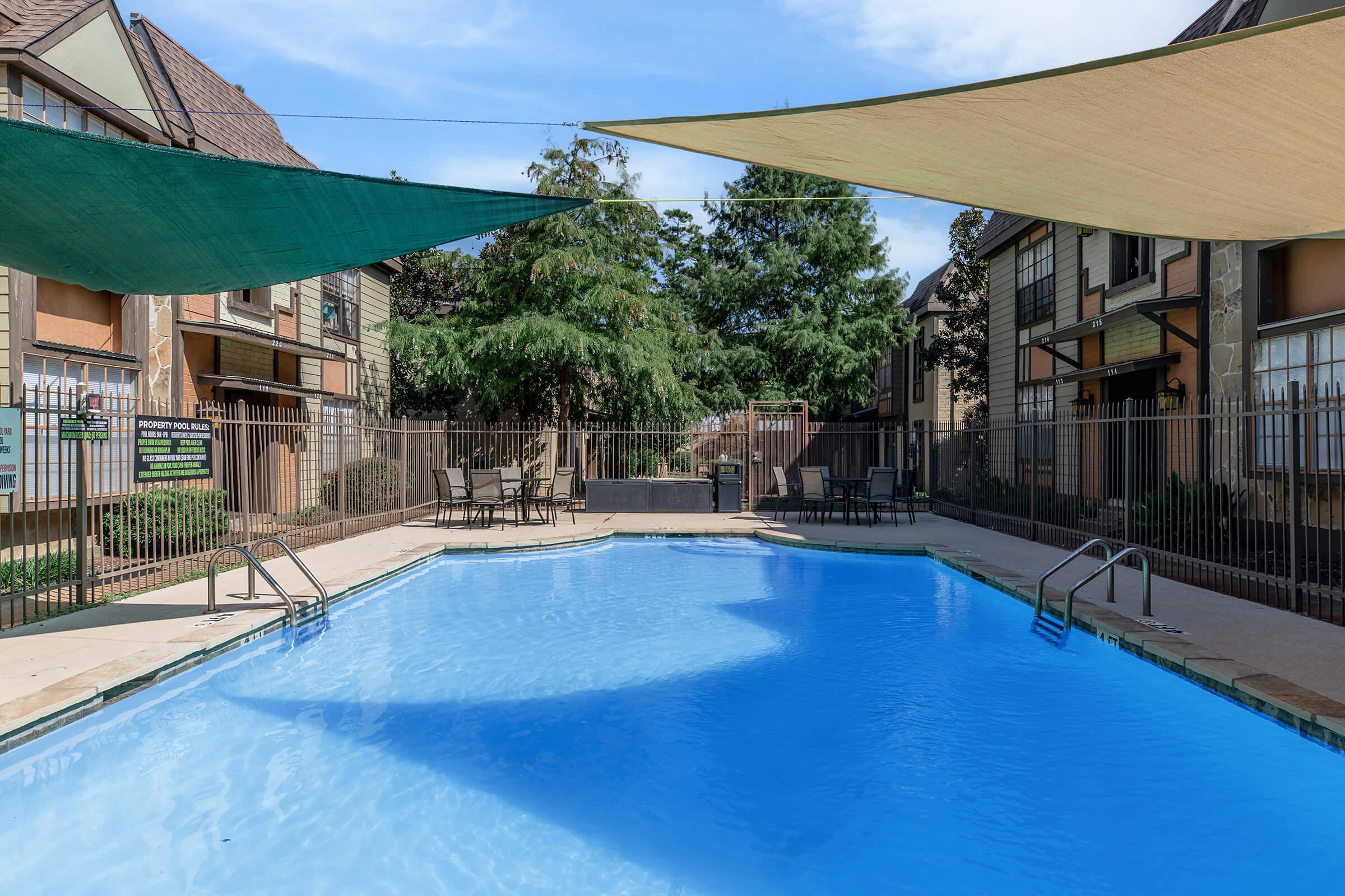 A swimming pool surrounded by green trees and apartment buildings. The pool is partially shaded by large canopies, and there are lounge chairs and tables along the poolside. The sky is clear, creating a bright and inviting atmosphere.