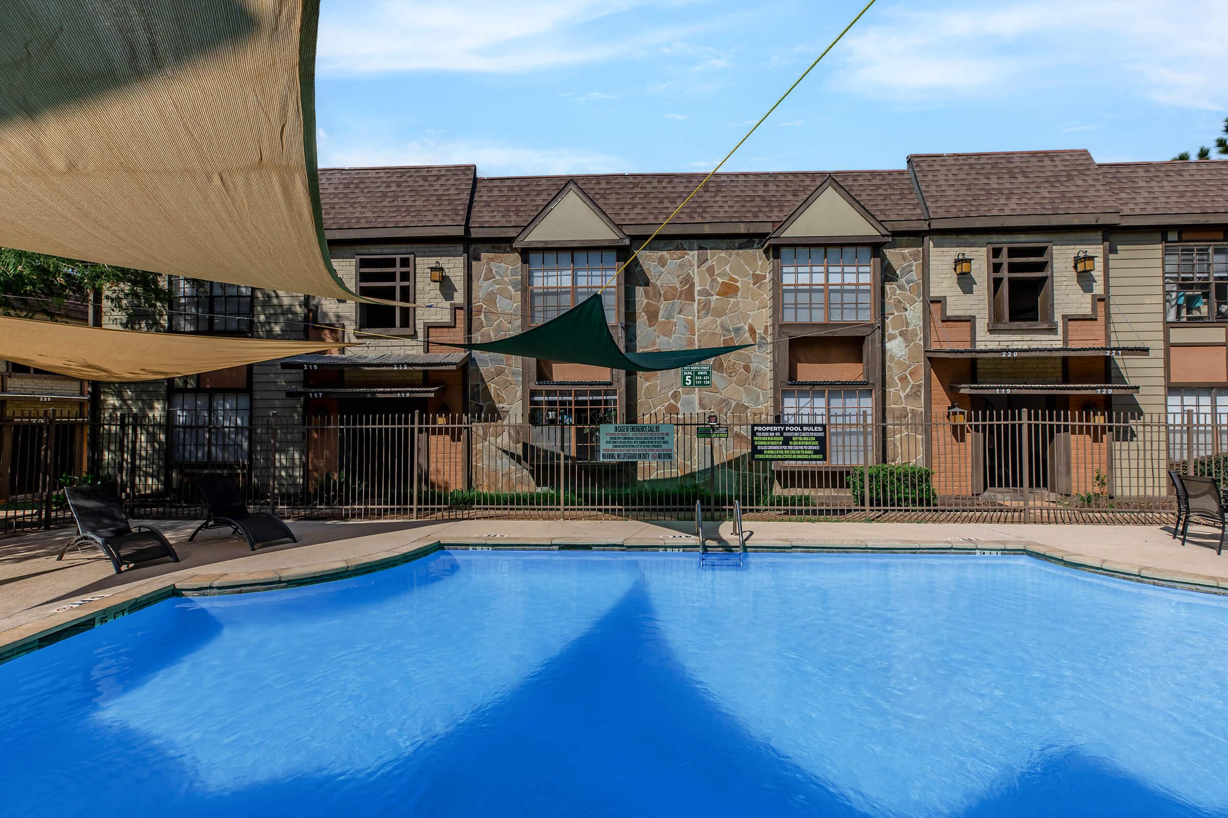 A clear blue swimming pool surrounded by a tan concrete deck and black lounge chairs. The background features a multi-story building with a stone façade and large windows, partially covered by green shade sails to provide outdoor comfort. The scene is serene with a bright blue sky overhead.