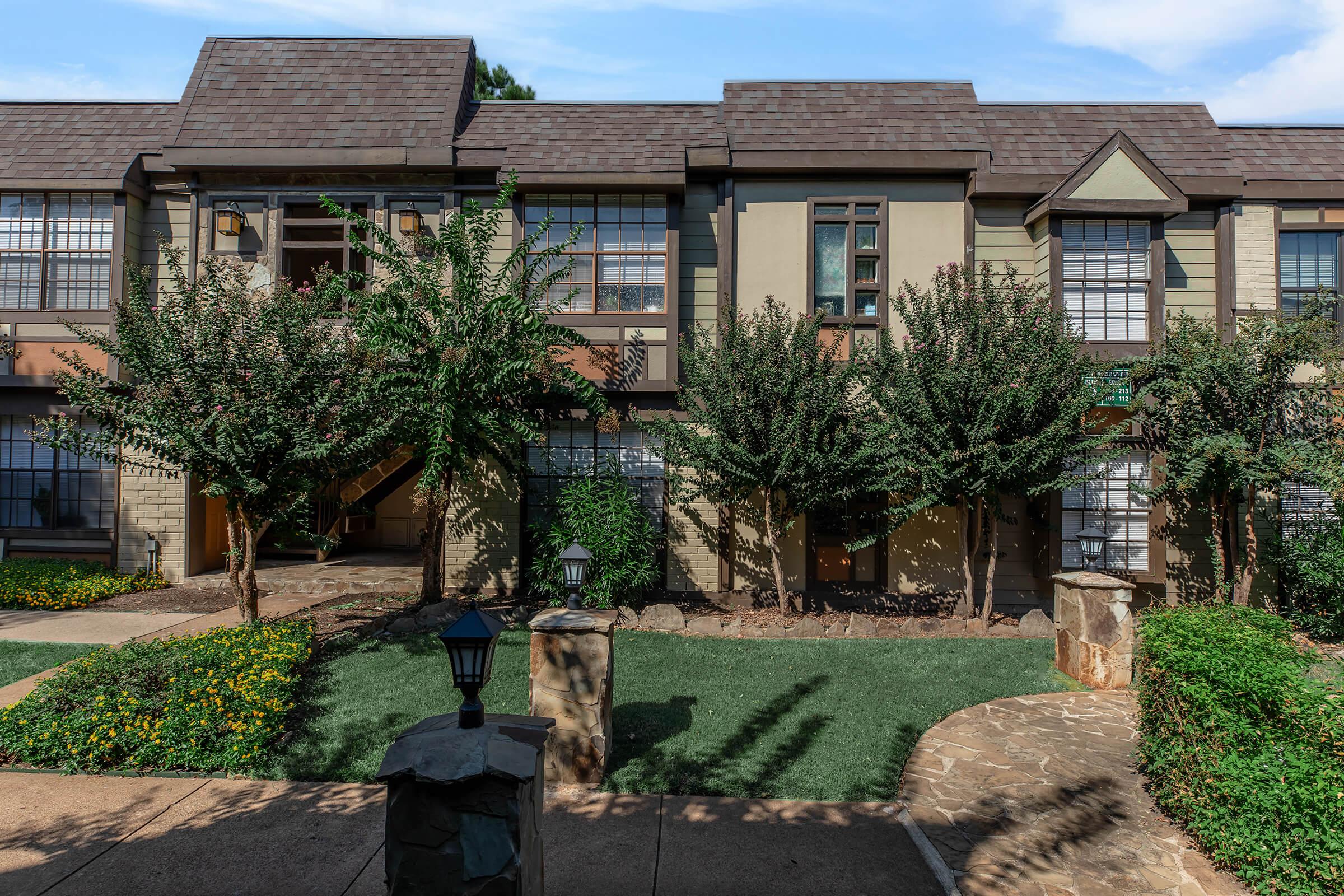 A view of a multi-unit residential building with a mix of green and beige siding. The building features multiple windows and is surrounded by landscaped areas with trees and shrubs. A stone walkway leads to the entrance, and a decorative lantern is visible in the foreground. Blue sky in the background.