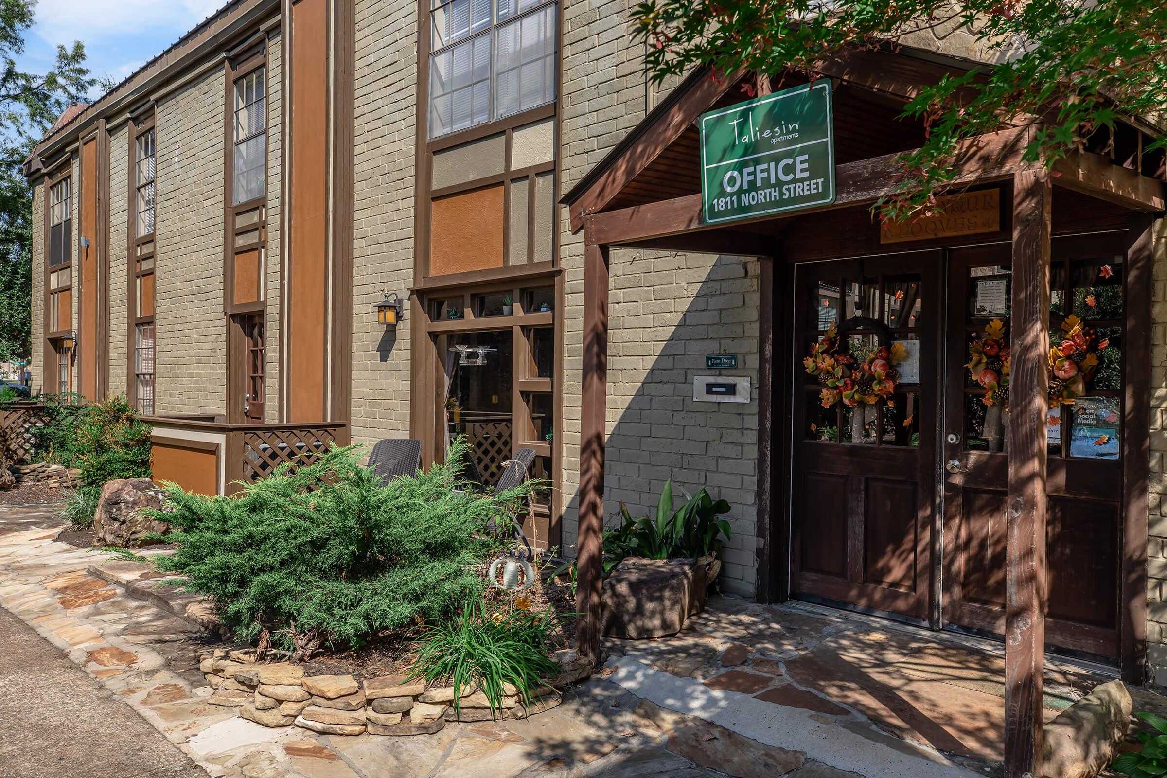 A rustic office building with a wooden entrance door adorned with seasonal decorations. The exterior features a mix of brown and green paint, large windows, and a sign reading "Office" with the address "5811 North Street." Lush greenery and a stone path lead to the entrance.