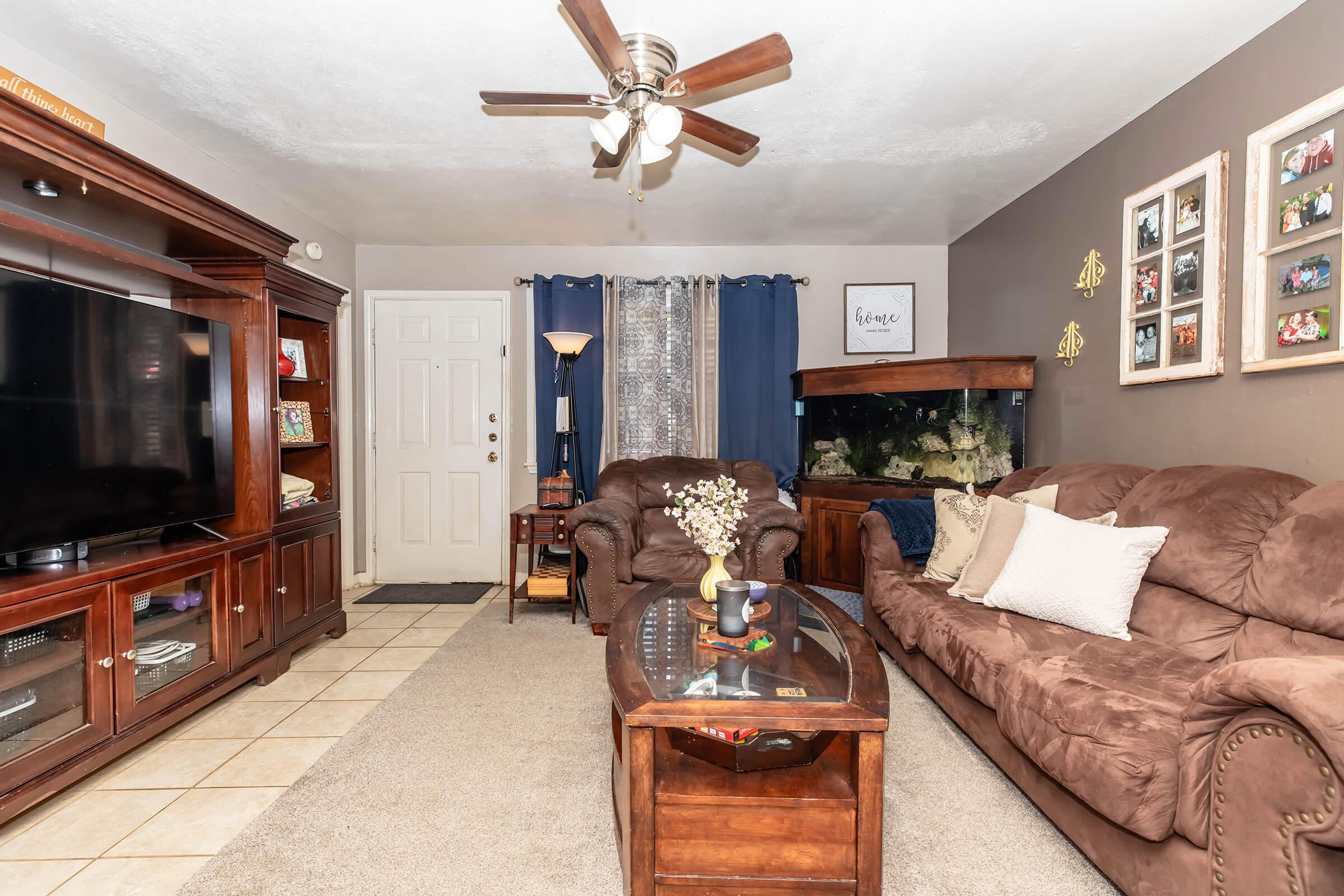 A cozy living room featuring a brown leather couch, a wooden coffee table, and a large TV unit. There's a wall-mounted fish tank, decorative photos framed on the wall, a standing lamp, and a door leading outside. The room has beige walls, a light beige carpet, and blue curtains by the window, creating a welcoming atmosphere.