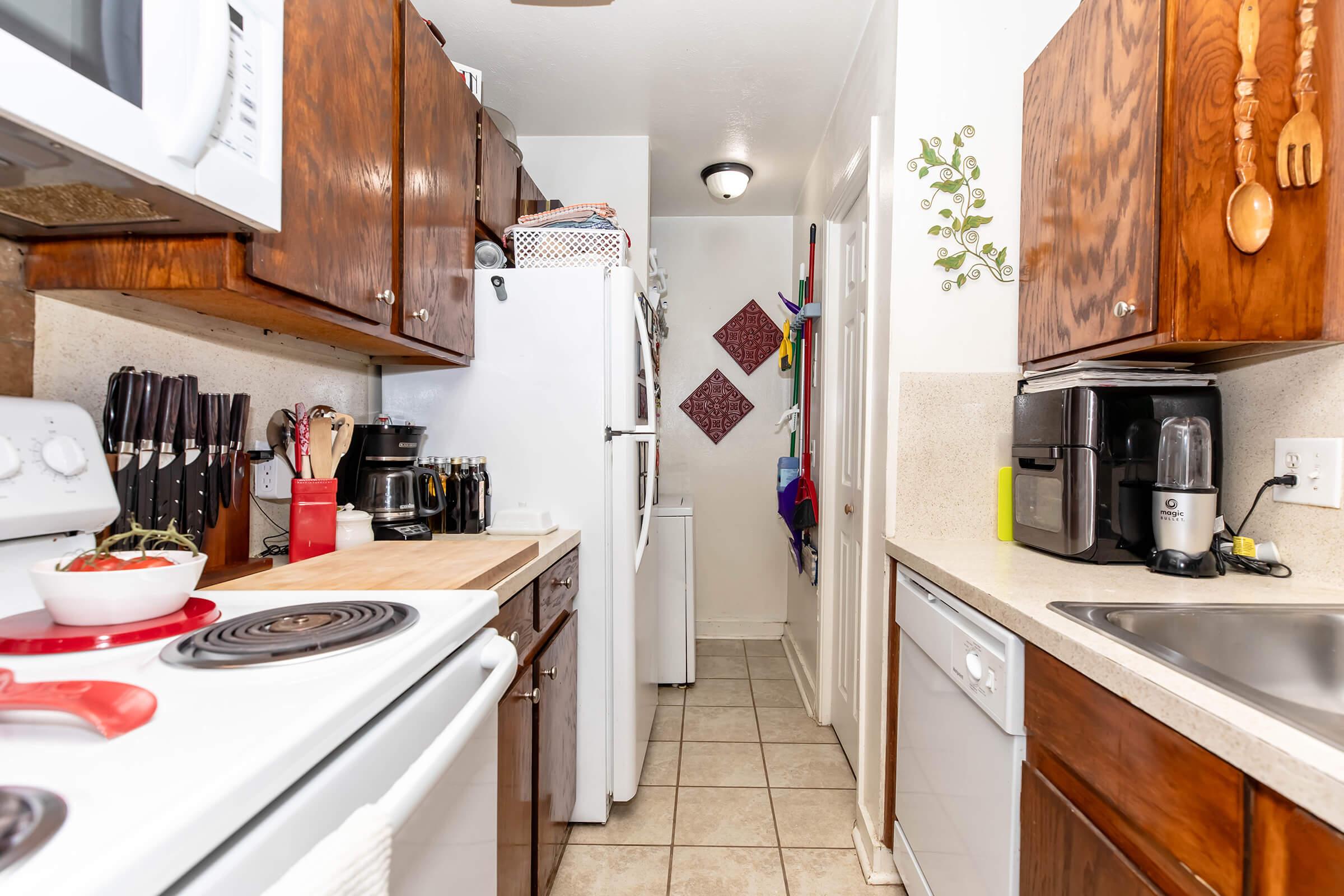 A narrow kitchen featuring wooden cabinets and white appliances. There's a stove, countertop, and a sink on one side. Against the opposite wall, a refrigerator and a closet door are visible. The space is well-organized, with utensils displayed and colorful items hanging on the wall, creating a warm, inviting atmosphere.