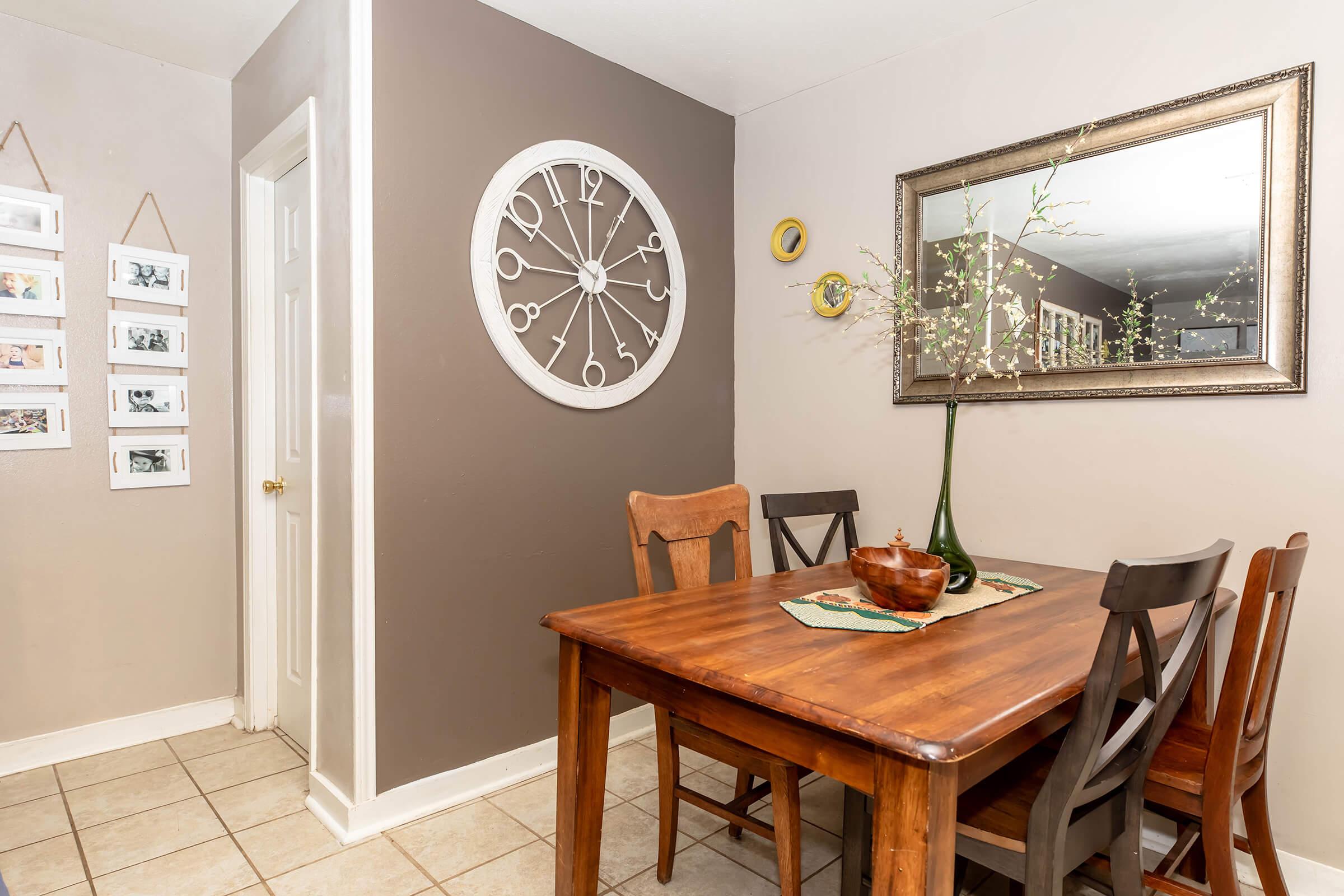 A cozy dining area featuring a wooden table with four chairs, a decorative bowl centerpiece, and a tall vase with branches. The wall is adorned with a large round clock and a mirror, while framed pictures are displayed on the adjacent wall. The flooring is tiled, creating a warm and inviting atmosphere.