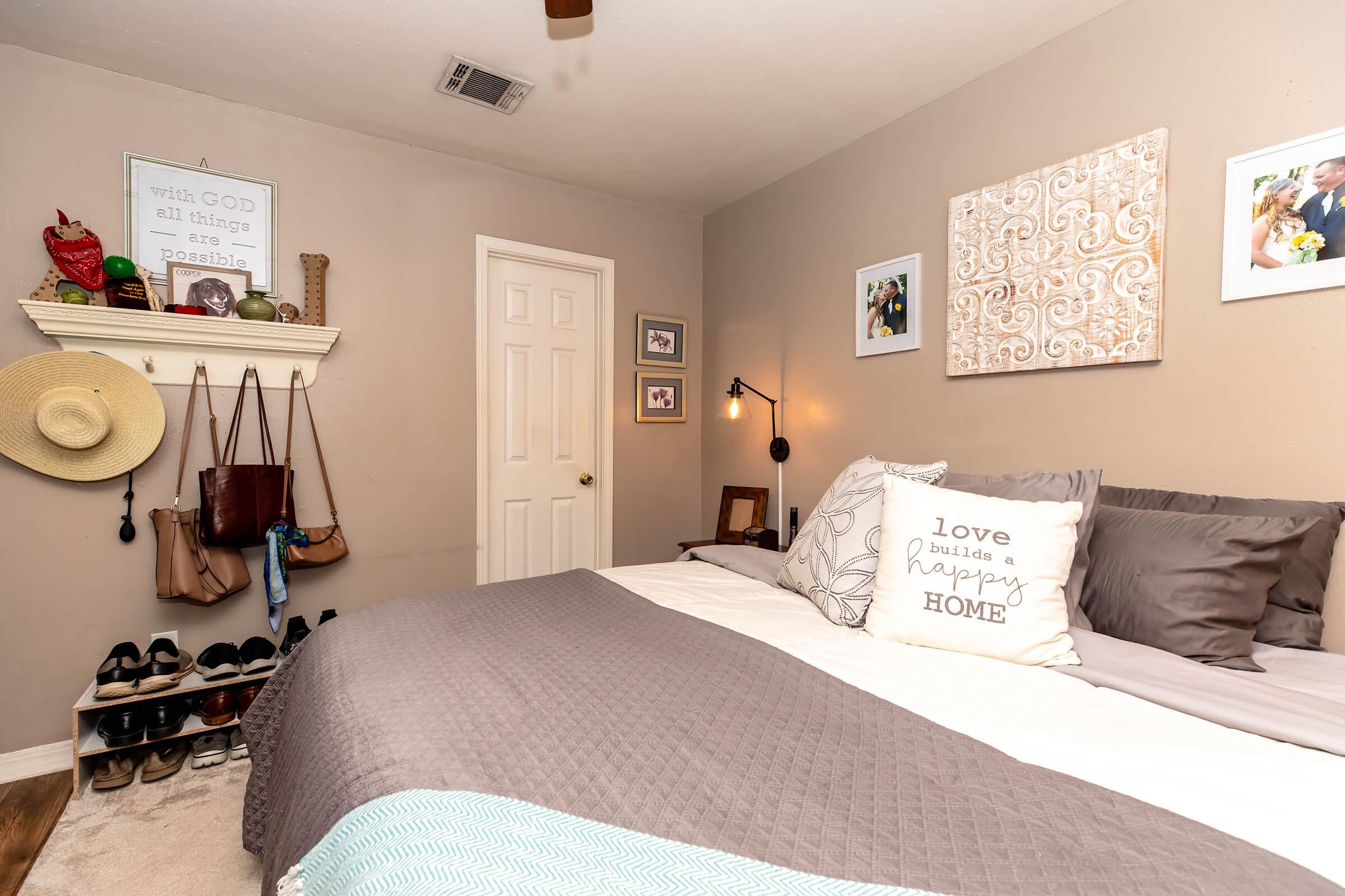 A cozy bedroom featuring a bed with gray and white bedding, adorned with decorative pillows. The walls are painted in a soft neutral color and decorated with framed photos and artwork. A shelf holds bags and accessories, and a pair of stylish shoes is neatly arranged on the floor. A light fixture provides warm illumination.