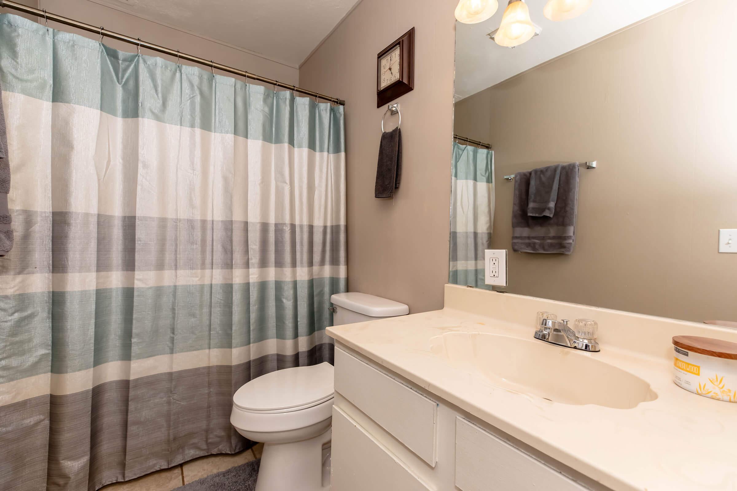 A clean bathroom featuring a light-colored countertop with a sink, a toilet, and a shower with light blue and gray striped curtains. The walls are painted a neutral beige, and there is a rectangular mirror above the sink. A hand towel hangs on the wall, and a small decorative item is on the counter.