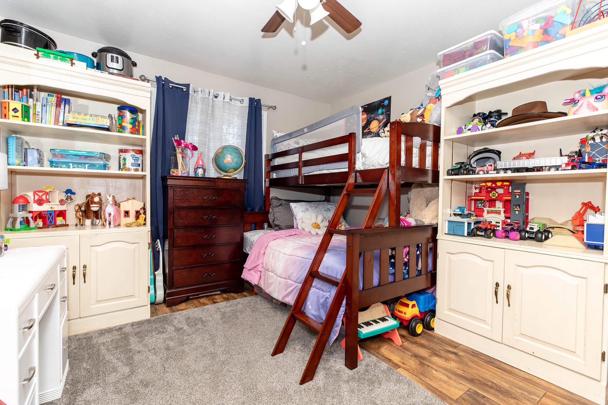 A cozy children's bedroom featuring a wooden bunk bed with colorful bedding, shelves filled with toys and books, a dark wooden dresser, and a soft gray carpet. Bright natural light streams through the window, creating a warm and inviting atmosphere.