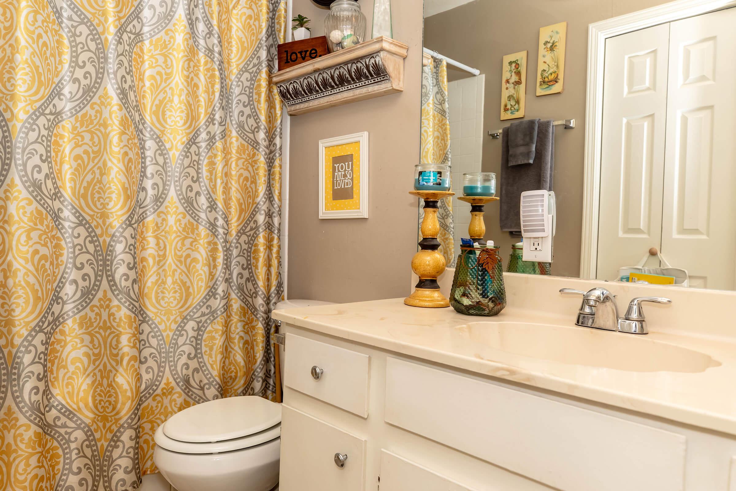 A neatly arranged bathroom featuring a light-colored vanity with a marble countertop, a modern sink and faucet, and a white toilet. The space is accented by a decorative shower curtain with a yellow and gray pattern and a small shelf displaying decor. Blue candles and framed art add color and warmth to the room.