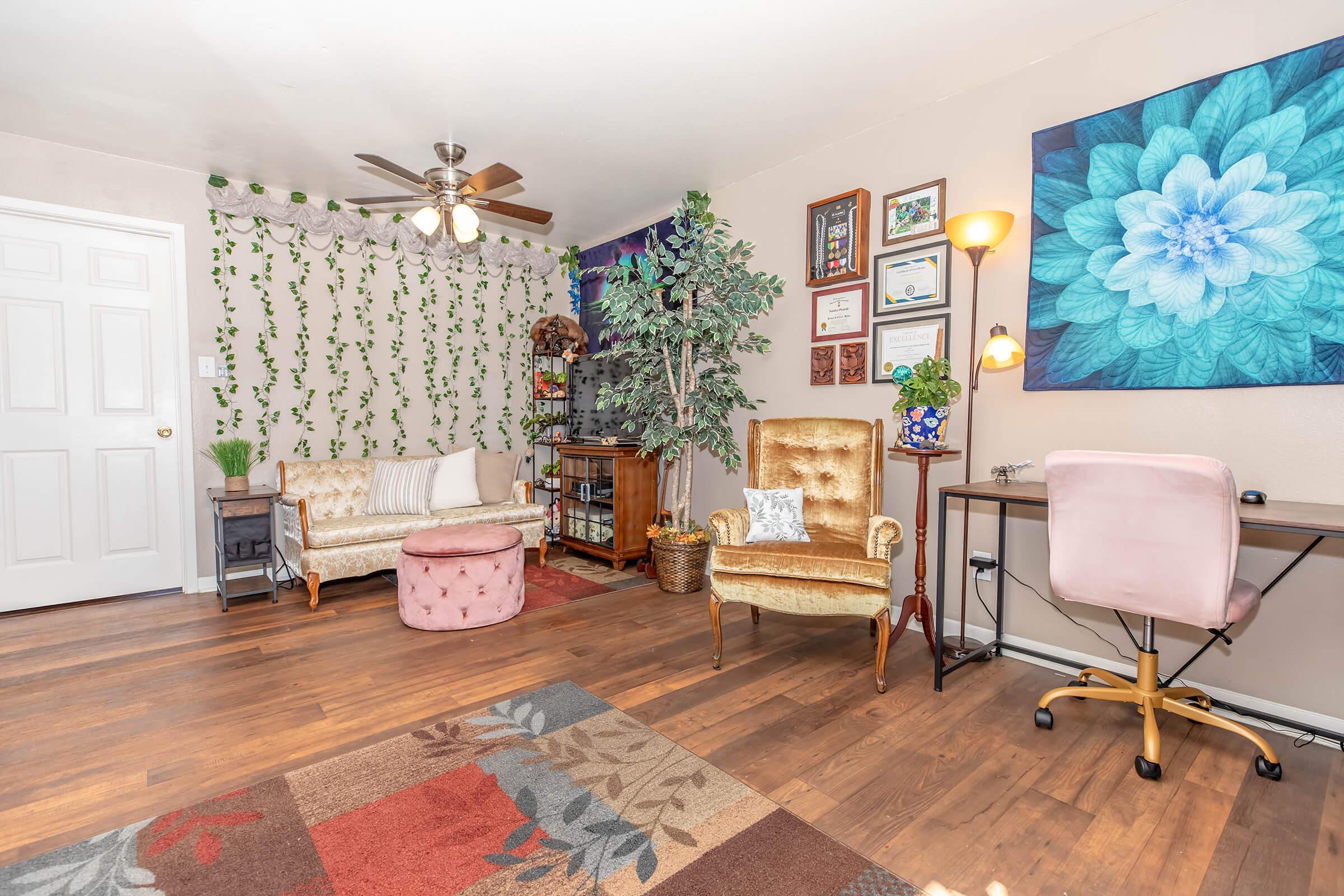 Cozy living room featuring a combination of vintage and modern furniture. Includes a golden armchair, a pink ottoman, a wooden bookshelf with plants, and a pink desk with a lamp. Walls are adorned with certificates and a floral artwork. The floor has a patterned rug, enhancing the inviting atmosphere.