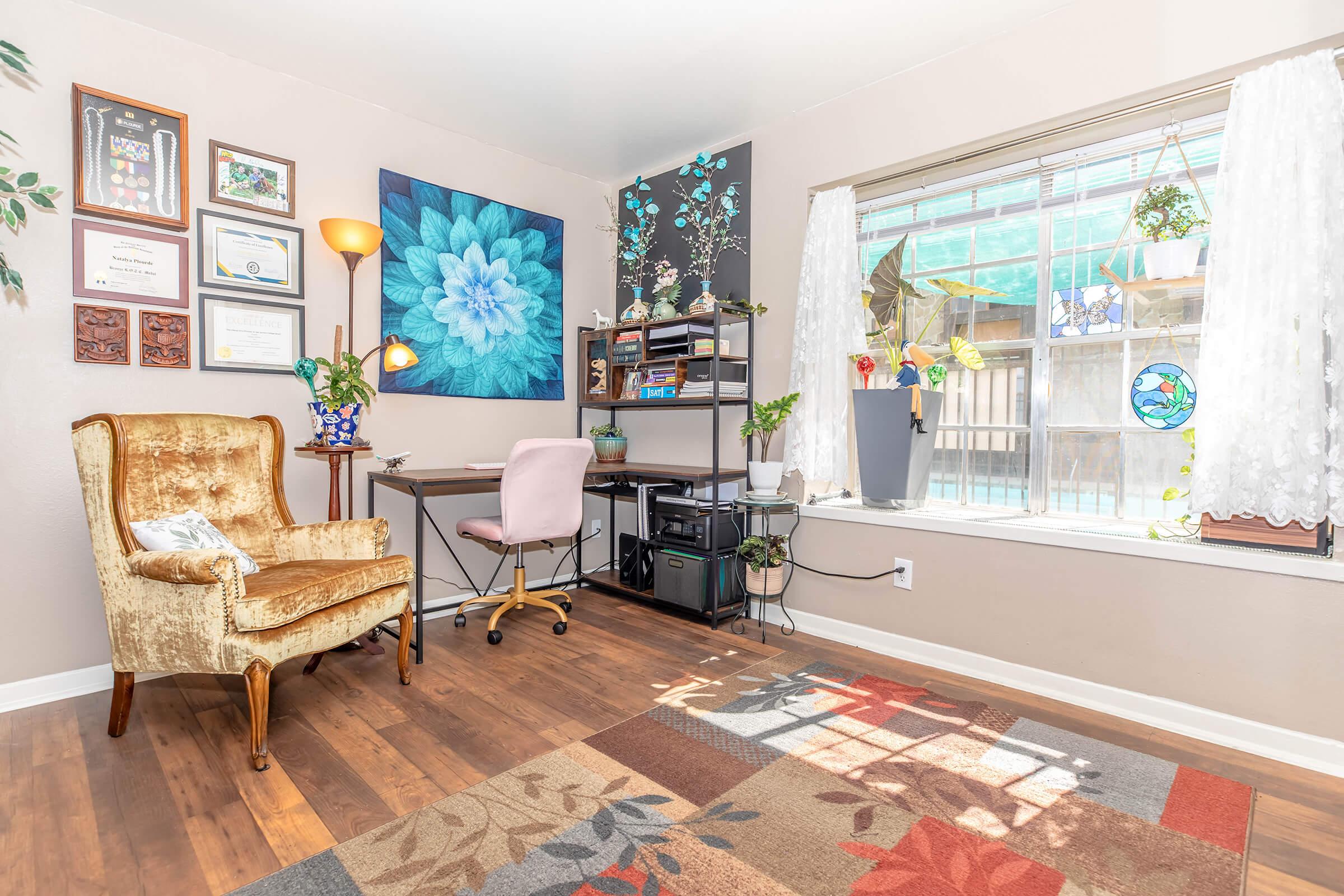 A cozy home office featuring a gold armchair and a light pink desk with a computer. The wall displays colorful artwork and framed certificates. A large window lets in natural light, and potted plants decorate the space. The floor is covered with a patterned rug, adding warmth to the room.