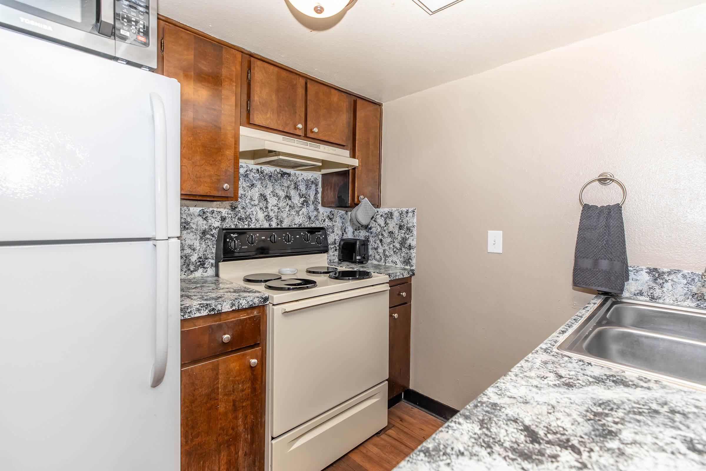 A small kitchen featuring dark wooden cabinets, a white refrigerator, and an off-white stove with a hood. The countertops are a marbled gray pattern, and there is a double sink with a gray towel hanging on a rack. A microwave is mounted above the stove. The overall atmosphere is clean and functional.