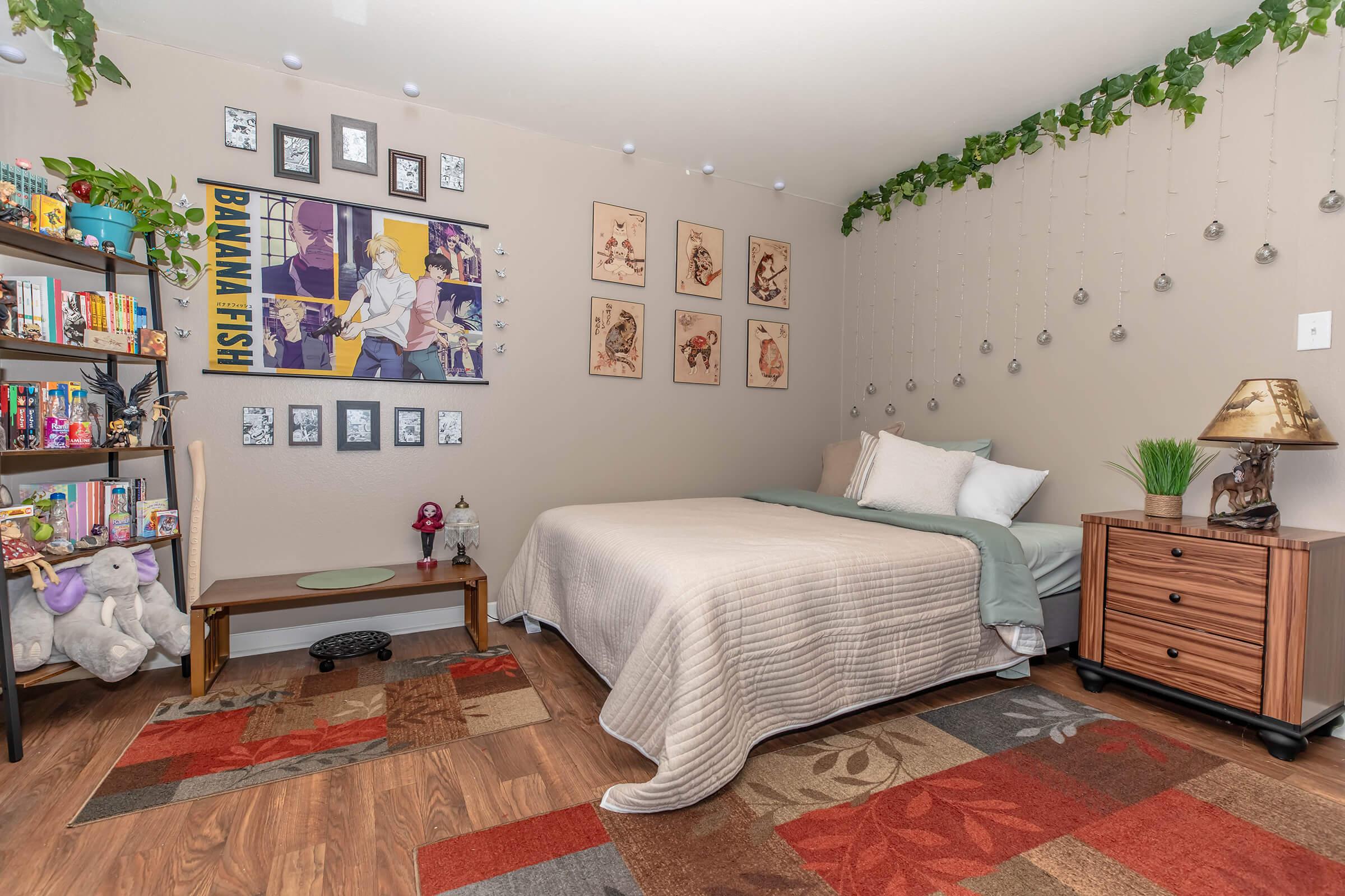 A cozy bedroom featuring a neatly made bed with light bedding, a vibrant area rug, and decorative wall art, including anime posters. A wooden bookshelf filled with books and plush toys is on one side, while hanging plants and decorative elements add a touch of greenery. The room is warmly lit and inviting.