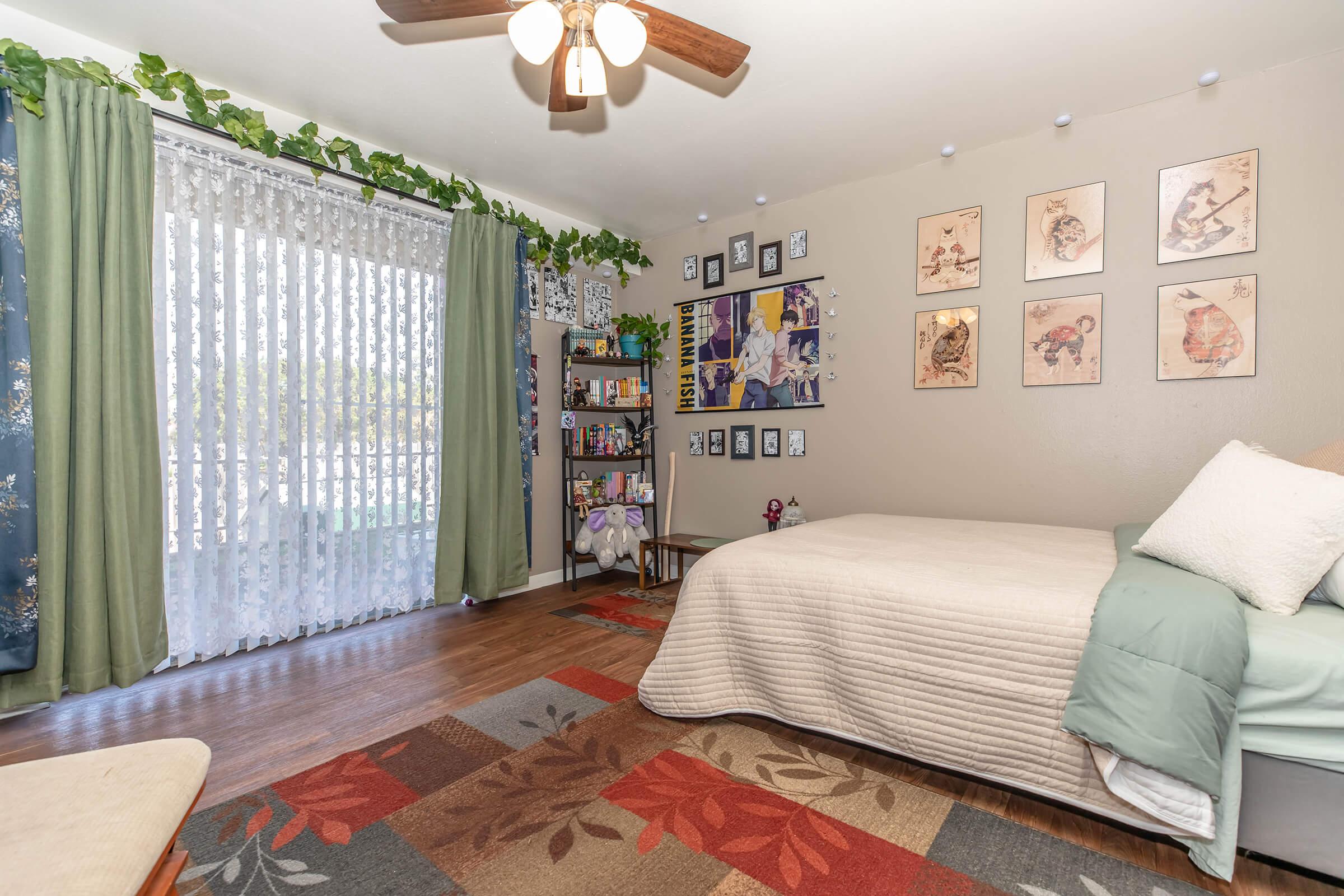 A cozy bedroom featuring a bed with a light-colored quilt, a decorative rug, and a bookshelf filled with various items. The room has green curtains, a flowy white curtain letting in natural light, and wall art displayed in frames. Potted plants add a touch of greenery to the space.