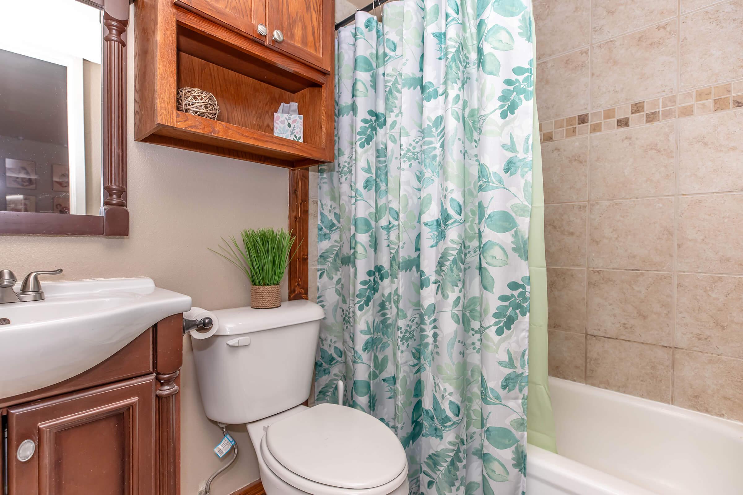 A small bathroom featuring a white sink and a toilet beside a bathtub. A green and white floral shower curtain partially covers the tub. Above the sink, there are wooden shelves with decorative items and a small potted plant on the countertop. The walls are tiled with light beige tiles.