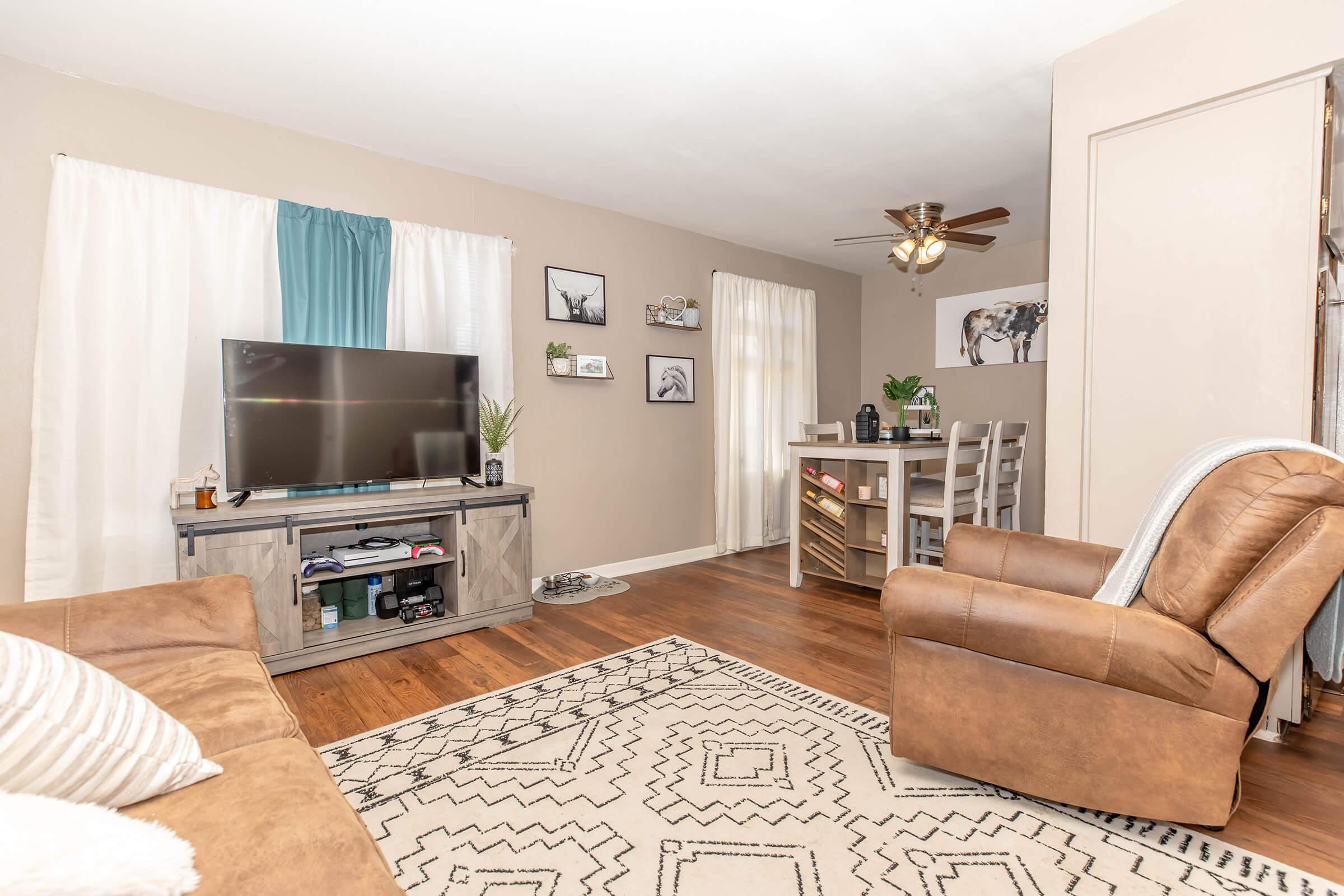 A cozy living room featuring a brown recliner, a light-colored sofa with decorative pillows, and a large flat-screen TV on a wooden console. The walls are adorned with framed art, and there's a dining area with a small table and chairs. Natural light filters in through sheer curtains, and a patterned rug adds warmth to the space.