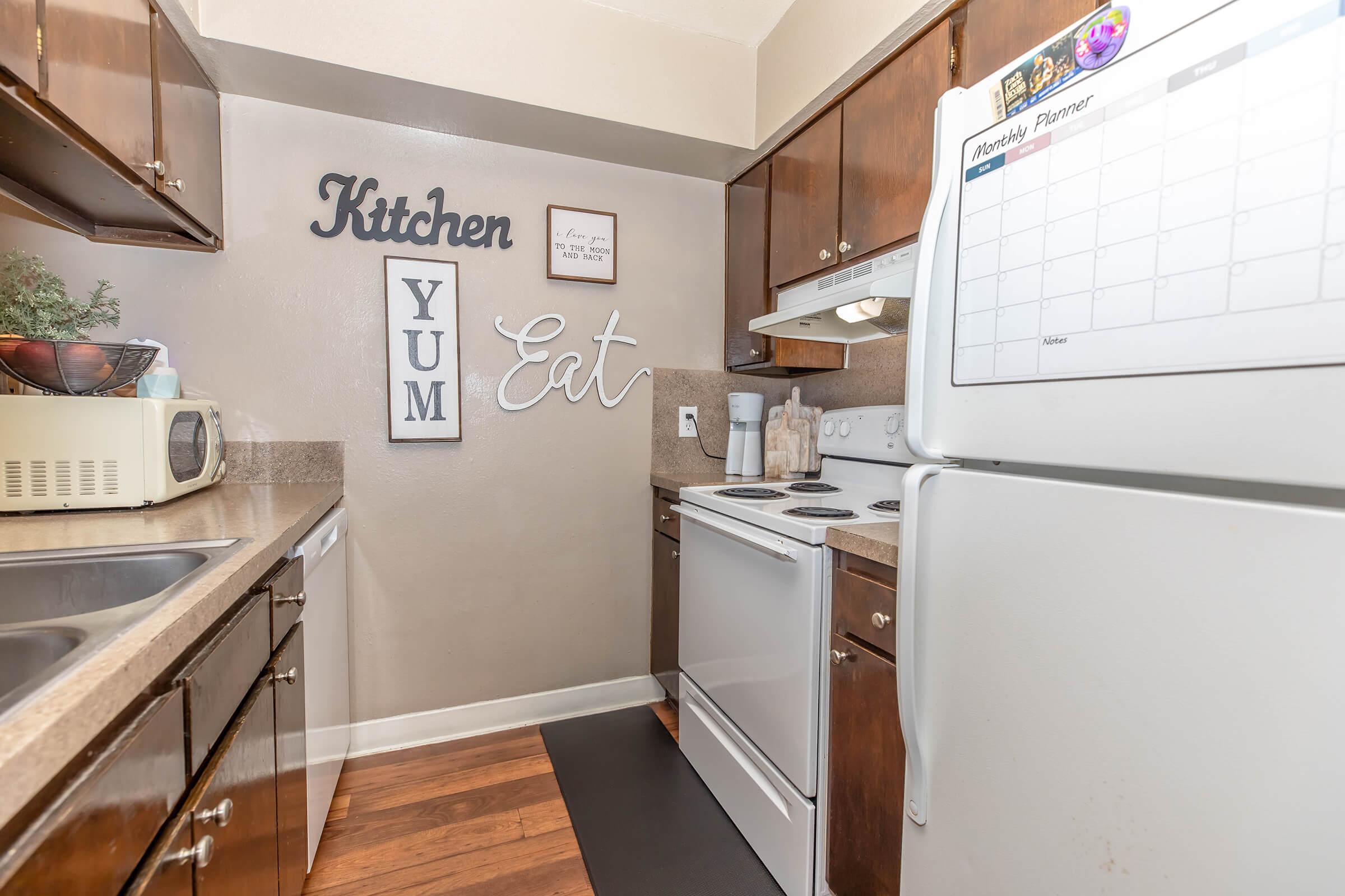 A cozy kitchen featuring wooden cabinets, a small sink, and a white stove. Decor includes a "Kitchen" sign, "Yum" and "Eat" lettering on the wall, and a refrigerator with a monthly planner on it. The floor has dark-colored vinyl, adding warmth to the space.