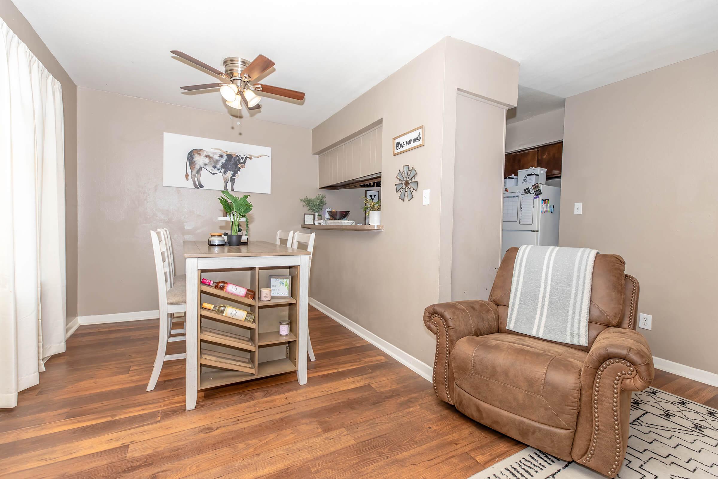 A cozy dining area featuring a small wooden table with four chairs, a brown recliner, and a ceiling fan. A cow-themed artwork hangs on the wall, while a partially visible kitchen area is seen in the background. Light curtains allow natural light to flood the room, enhancing the warm, inviting atmosphere.