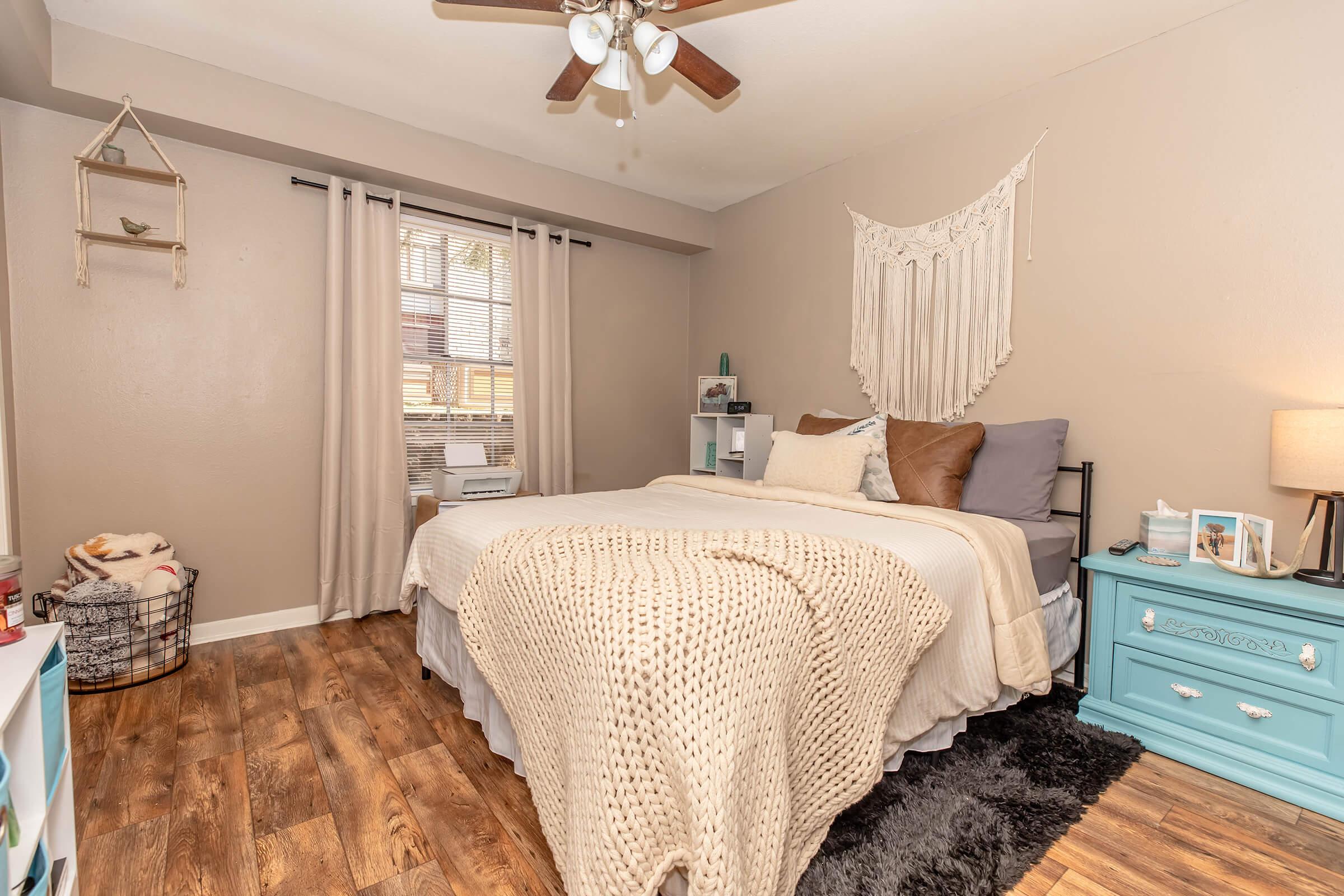 A cozy bedroom featuring a bed with neutral-toned bedding, a decorative wall hanging above, and a small bedside table with a lamp. The room has light brown walls, wooden flooring, and a window dressed with curtains. A small basket and decorative items add charm to the space.