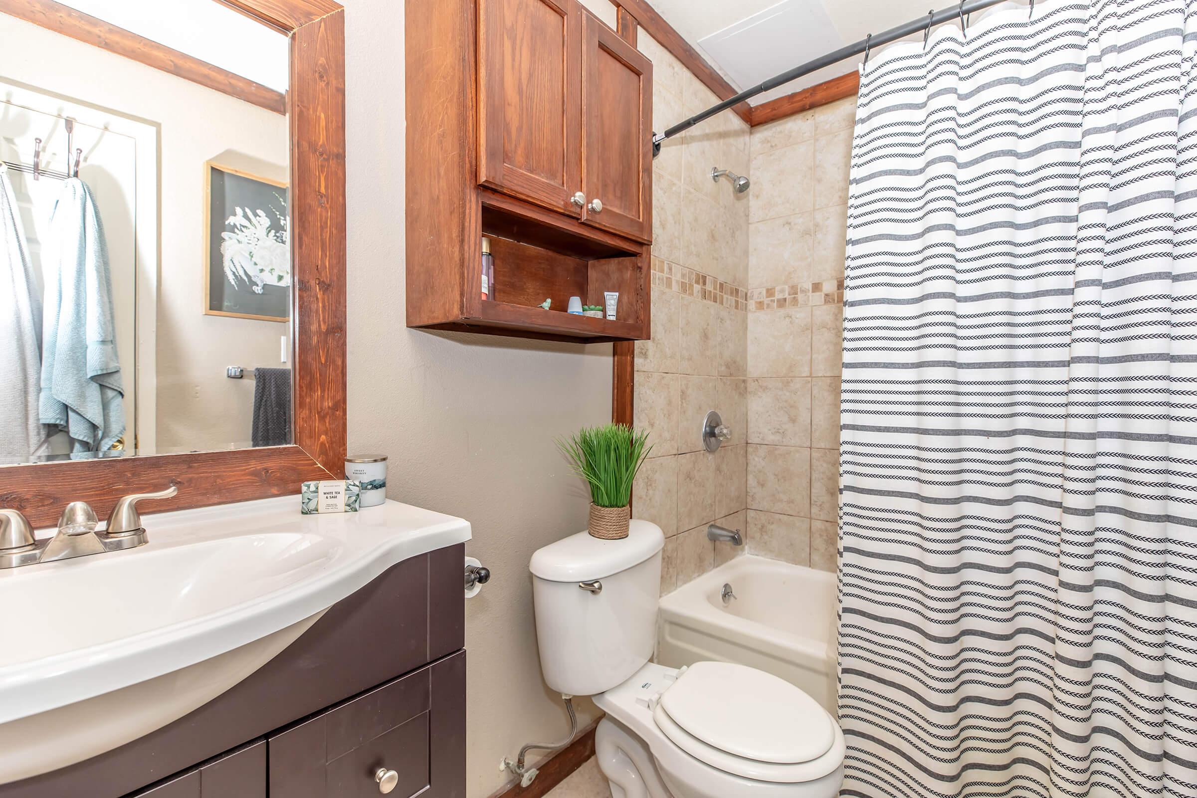 A well-lit bathroom featuring a white sink with a dark wood cabinet beneath, a white toilet, and a bathtub with a shower. The walls are tiled, and a striped shower curtain hangs in front of the tub. There is a small green plant on the toilet and a mirror above the sink with some toiletries on a shelf. Two towels hang nearby.