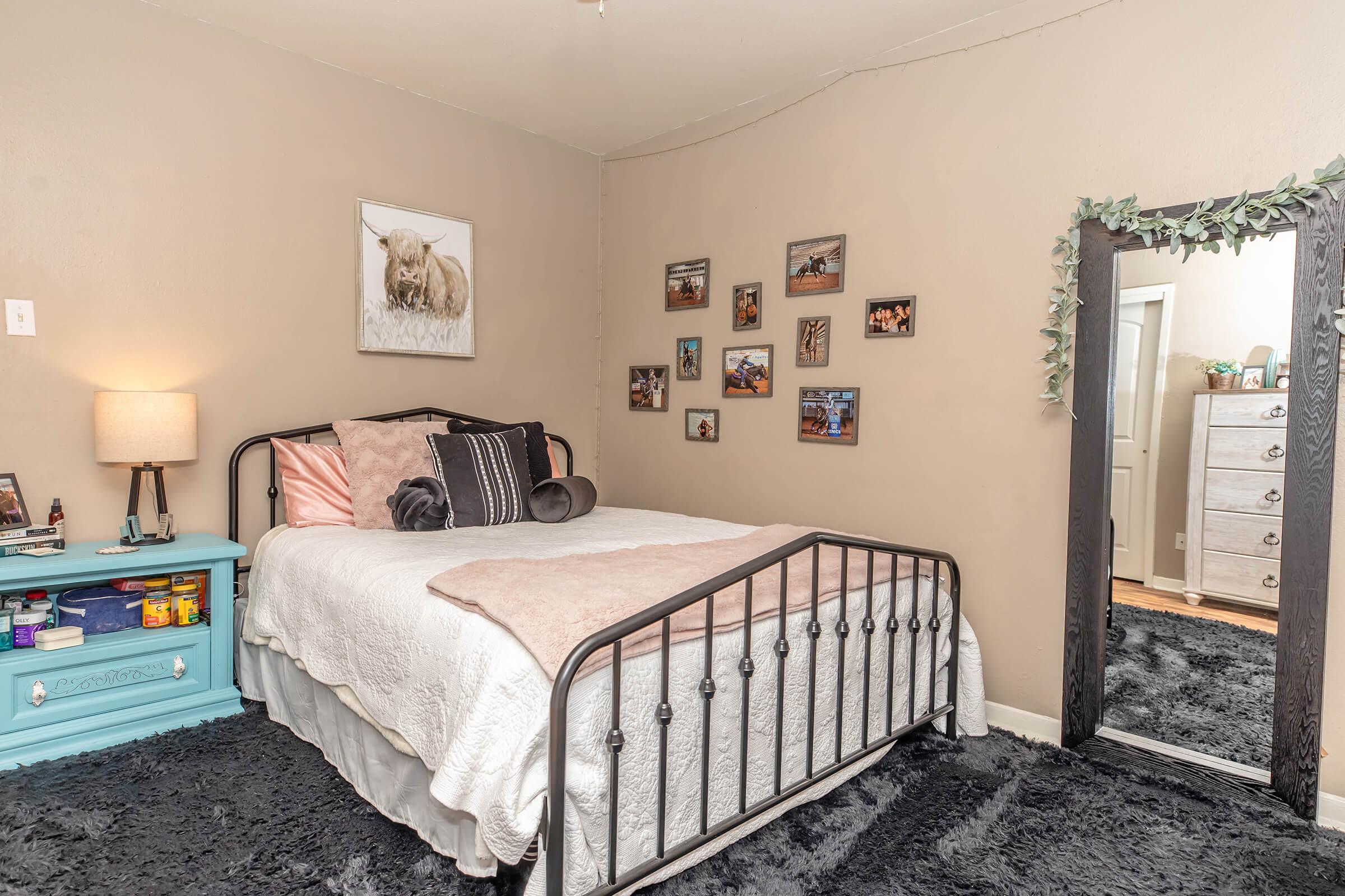 A cozy bedroom featuring a black metal bed with pink and white bedding, a nightstand with a lamp, and a vibrant blue dresser. The walls display framed photos, and there's a large mirror next to the bed. A soft black rug covers the floor, creating a warm and inviting atmosphere.