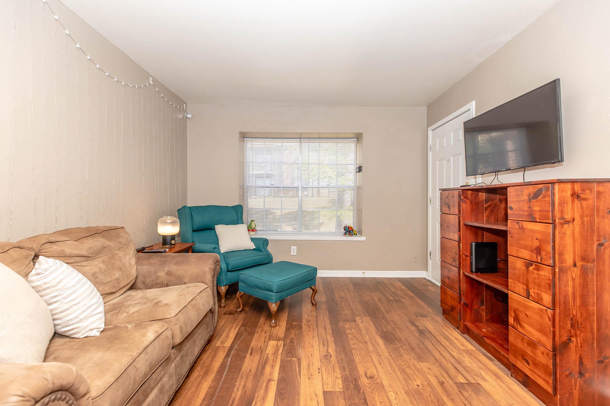 A cozy living room featuring a brown sofa and a teal armchair. There's a wooden TV stand with drawers and a television mounted on the wall. A window with blinds allows natural light to enter, and there are decorative items in the windowsill. The floor is made of light wood, creating a warm atmosphere.