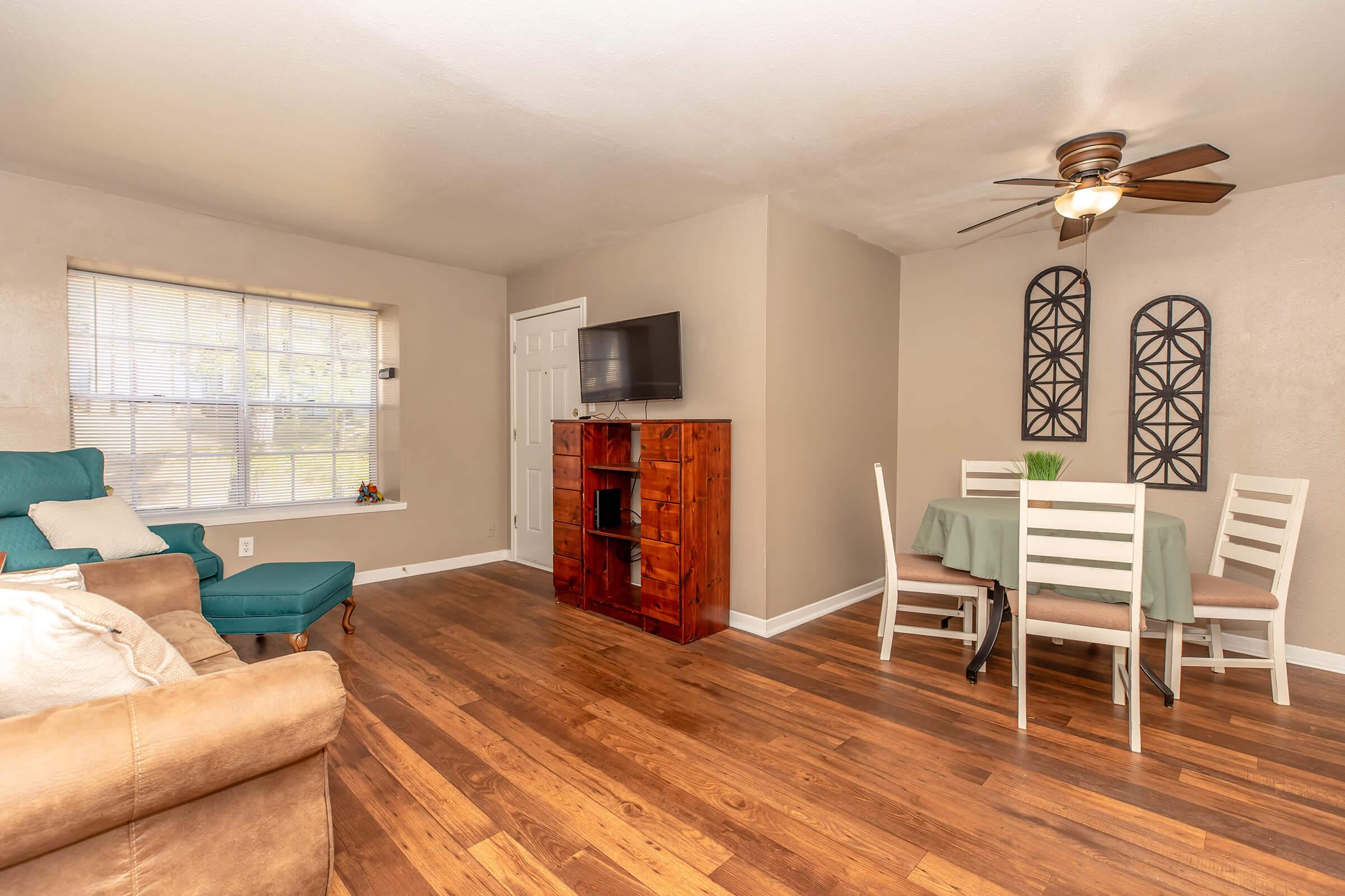 A cozy living area featuring a light-colored wall, wooden flooring, and natural light from a window. A blue armchair sits beside a beige sofa. A wooden cabinet and a small TV are mounted on the wall. A dining area with a green tablecloth, six white chairs, and decorative wall art completes the space.