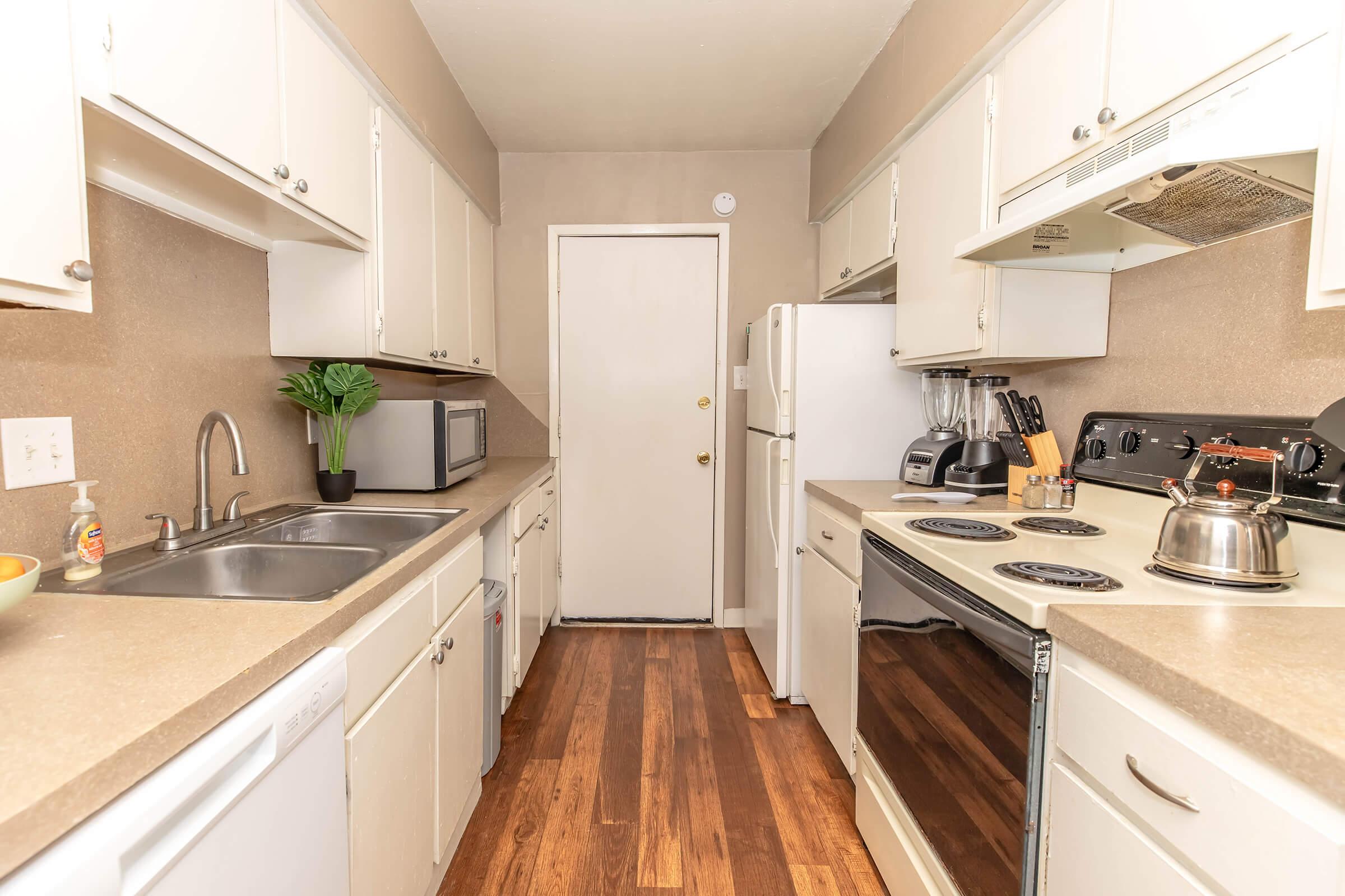 A modern kitchen featuring white cabinets, stainless steel appliances, and a double sink. The space includes a microwave, blender, and a kettle on the stove. Wooden flooring complements the beige walls, and a door leads to another area. A potted plant adds a touch of greenery to the countertop.