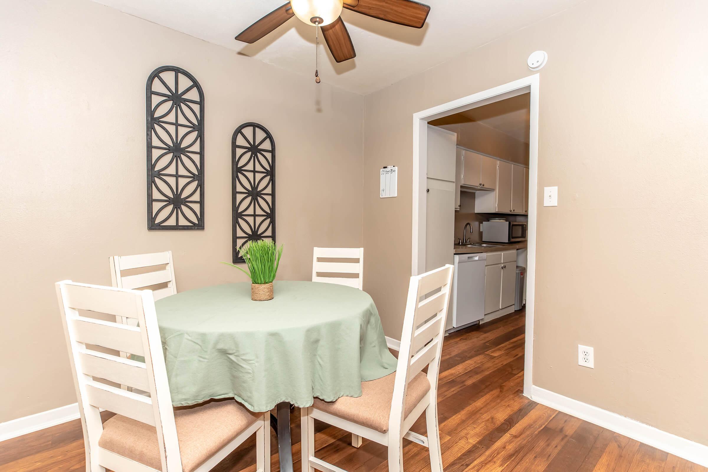 A cozy dining area featuring a round table covered with a green tablecloth, surrounded by four light-colored chairs. On the table is a small potted plant. The walls are painted in a warm tone, and decorative wall art is visible. A doorway leads to a kitchen with white cabinets in the background.