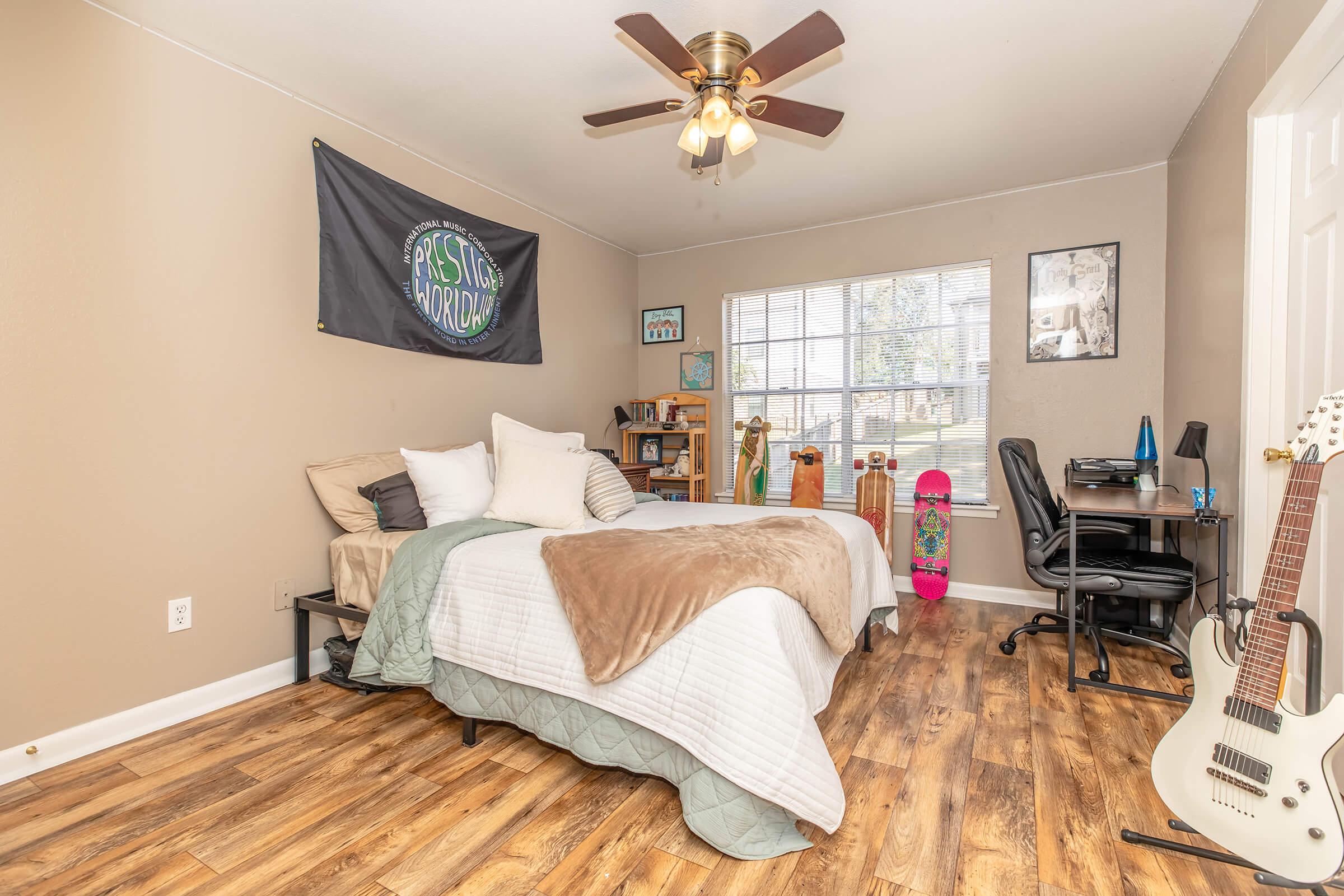 A cozy bedroom featuring a bed with a brown and white blanket, a ceiling fan, a black wall flag, and a large window. There’s a desk with a chair and computer, a bookshelf filled with items, and a few musical instruments. A skateboard decorates the room, along with wooden flooring.