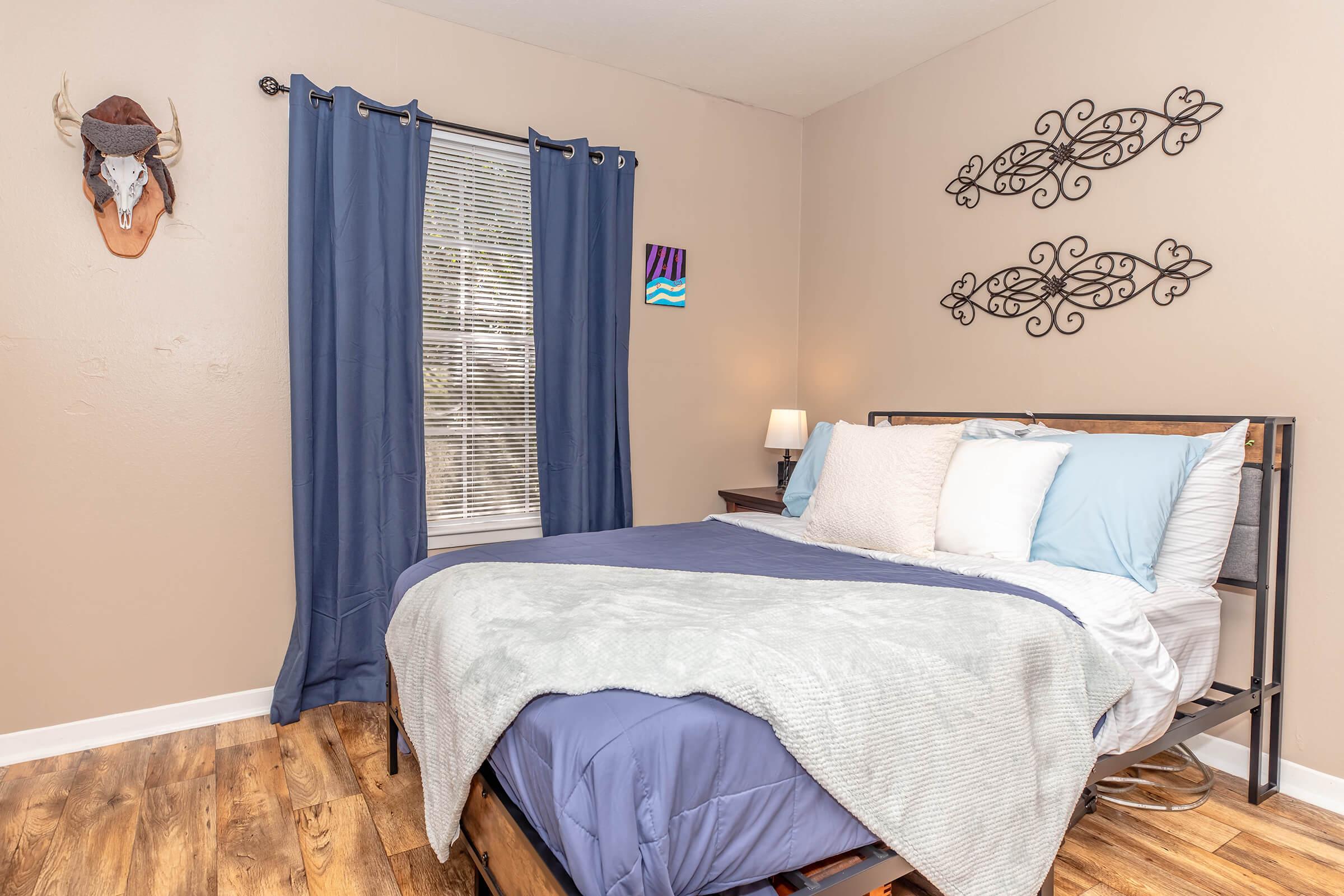 Cozy bedroom featuring a bed with blue and white bedding, decorative pillows, and a light gray throw. There's a window with blue curtains, a bedside lamp, and decorative wall art. An animal head mount adorns one wall, adding a rustic touch, while elegant metal wall decorations complete the warm ambiance.