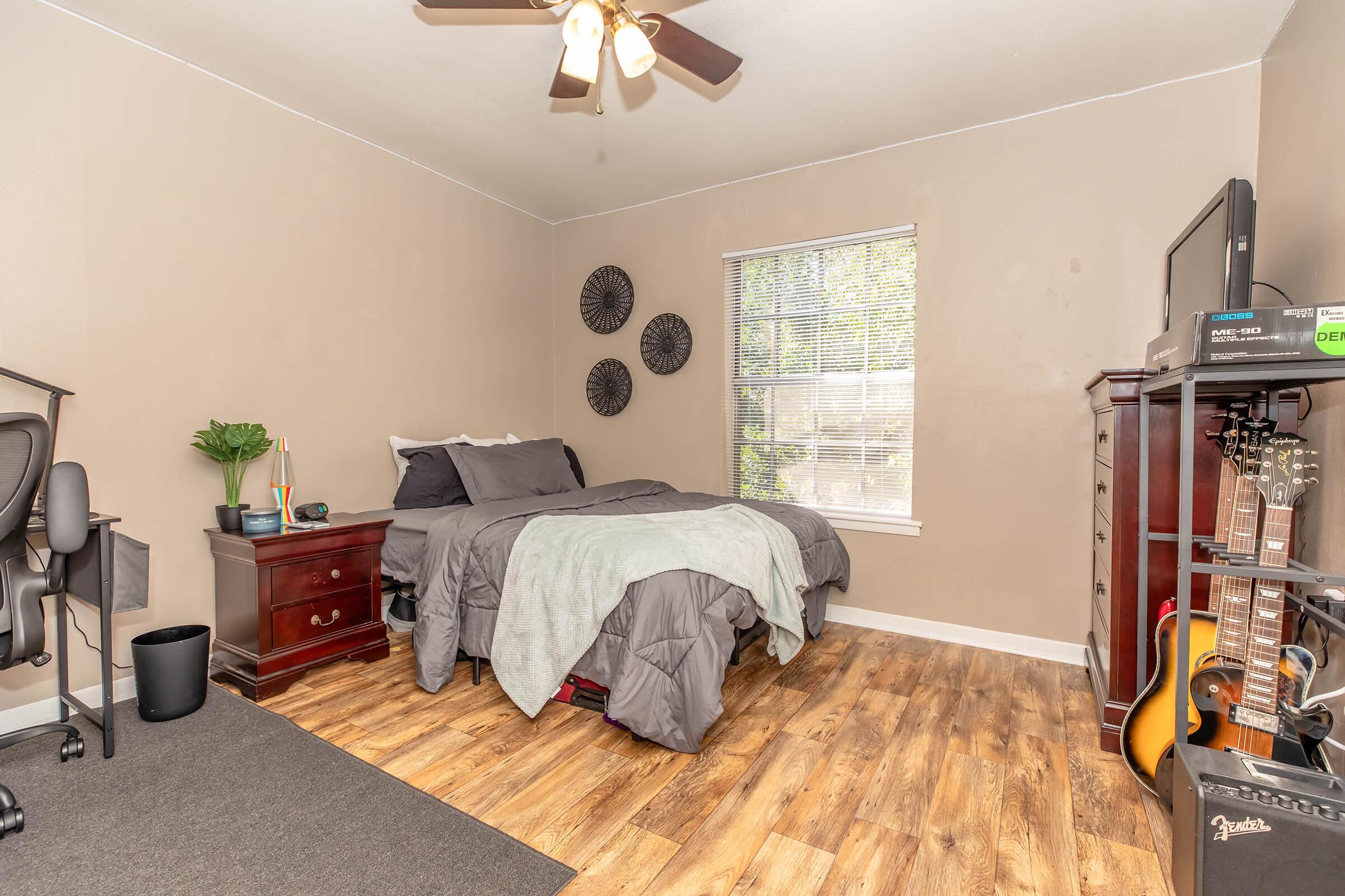 A cozy bedroom featuring a queen-sized bed with gray bedding, a nightstand, and a window with blinds. There is a black office chair in one corner, a small plant on the nightstand, and a dresser with a television. A guitar leans against the dresser, and the room has wood-like flooring with a dark rug.