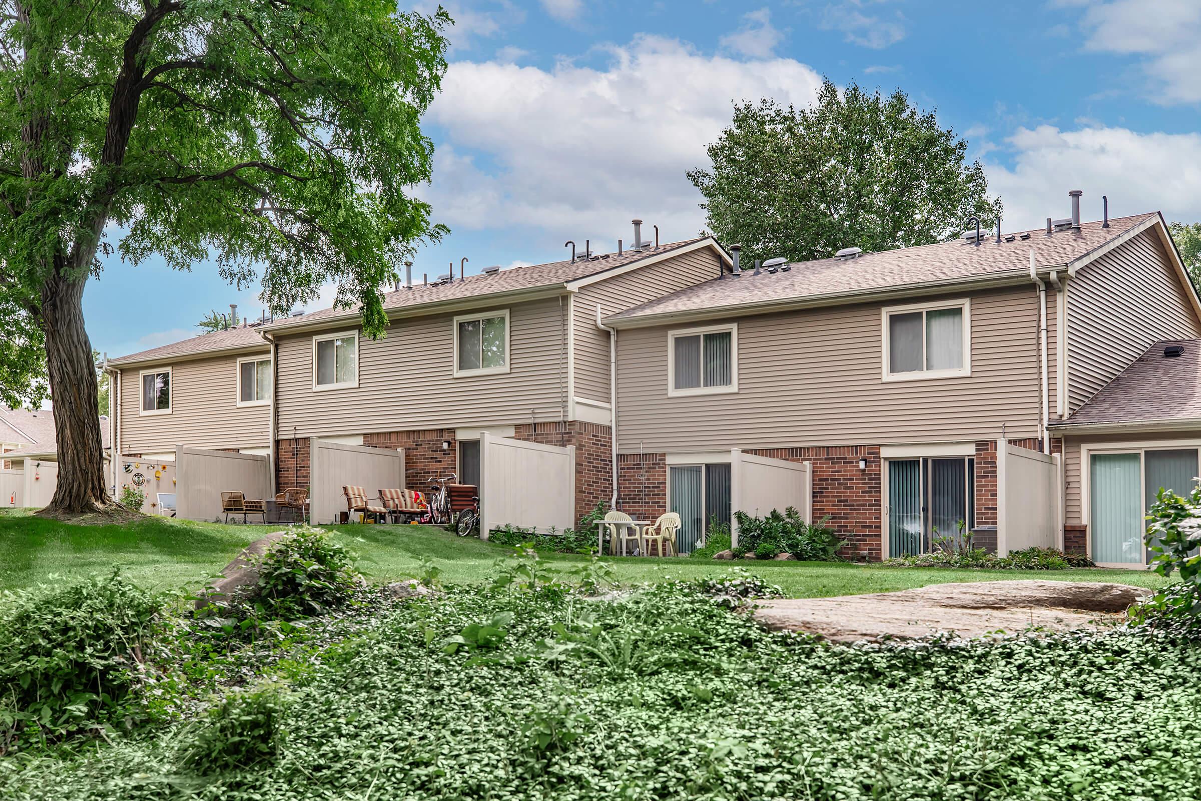 A row of two-story townhouses with beige siding and brick accents, surrounded by greenery. The scene includes a tree on the left and outdoor seating on a small patio in front of one of the units. The sky is partly cloudy, creating a serene suburban atmosphere.