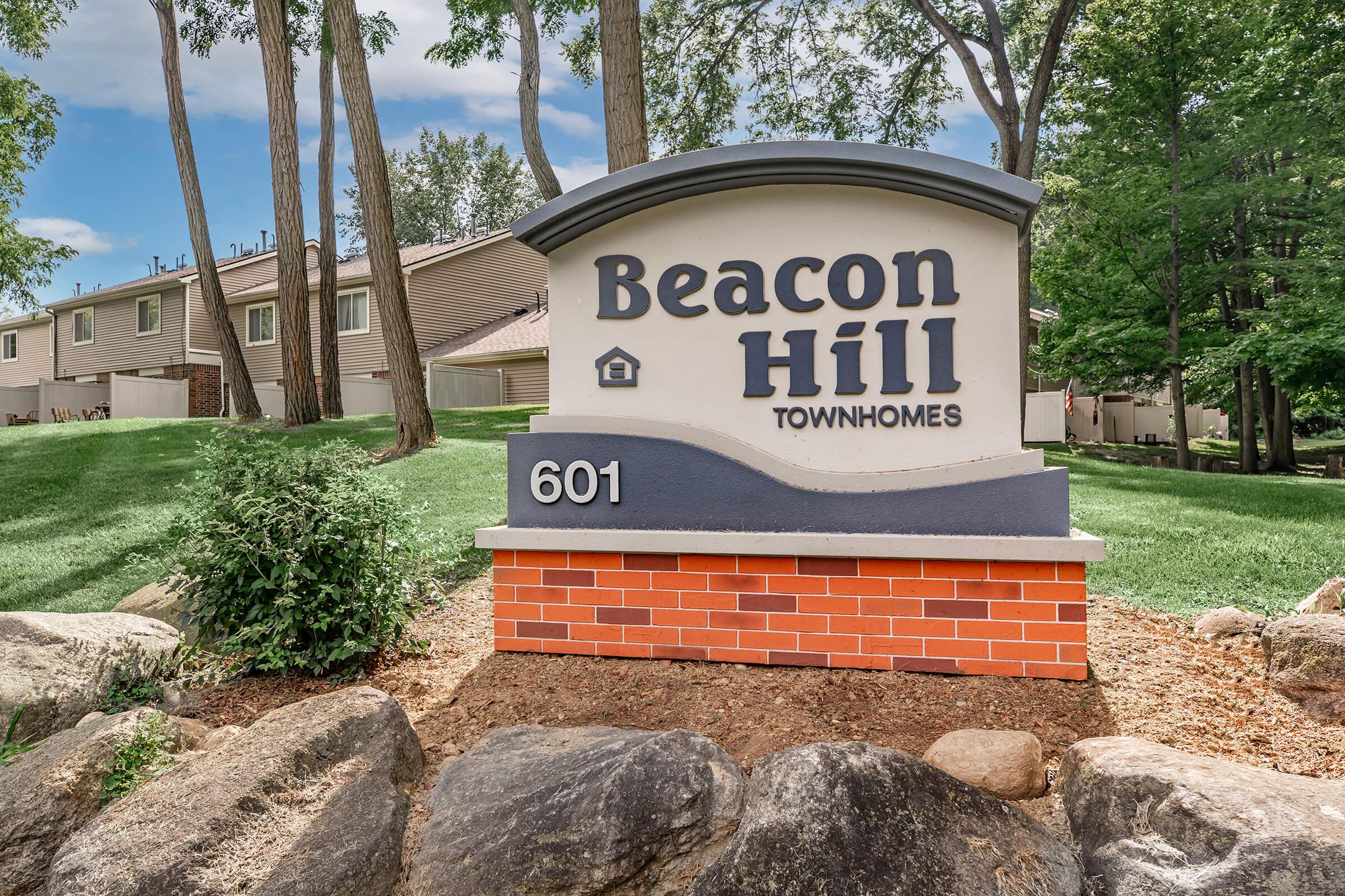 Sign for Beacon Hill Townhomes, featuring the name "Beacon Hill" prominently at the top, with "Townhomes" below. The sign is situated near lush greenery and rocks, with a background of trees and residential buildings. The address "601" is displayed at the bottom of the sign.