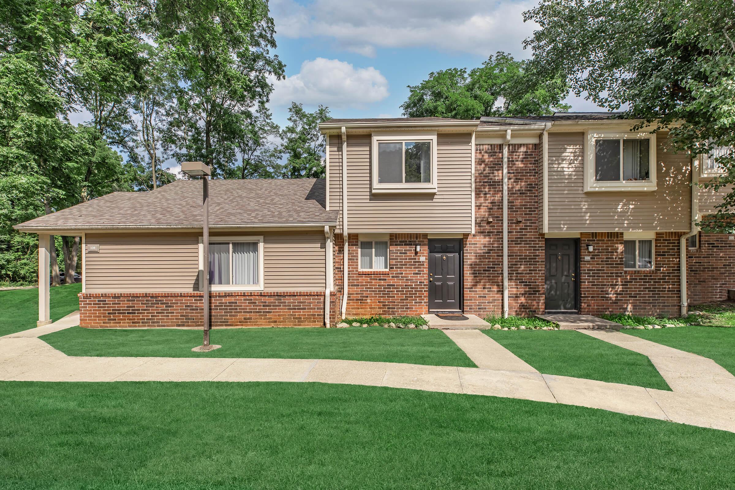 Two residential buildings with a mix of brick and beige siding, located in a green landscape. There are two doors on each building, black in color, and a pathway runs in front of them. The surrounding area features grass and trees under a partly cloudy sky.