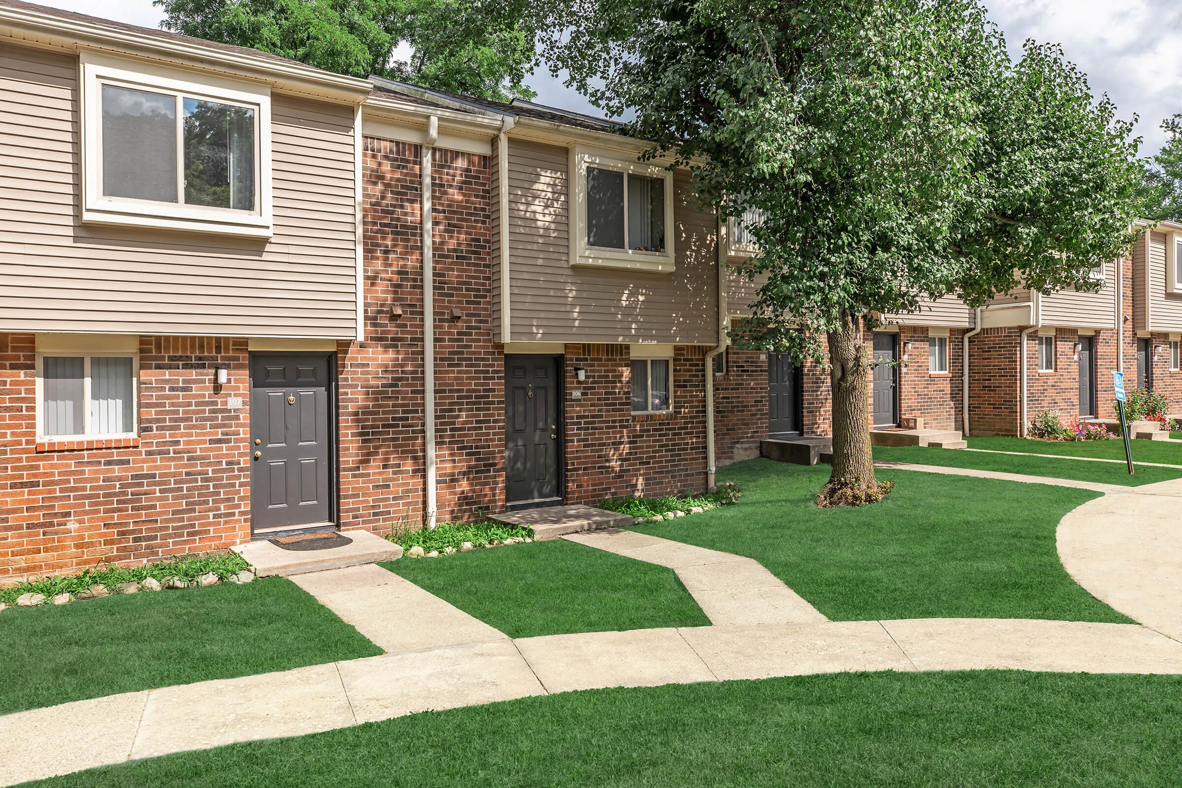 A row of two-story residential buildings featuring brick and siding exteriors. Each unit has a front door and small walkway, surrounded by well-maintained green lawns and trees. The scene is bright with a partly cloudy sky, creating a welcoming neighborhood atmosphere.
