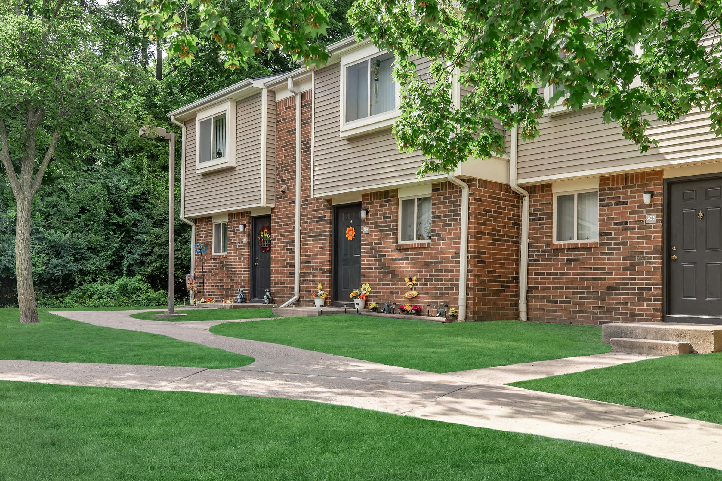 A well-maintained residential area featuring two townhouses made of brick. The front yards are landscaped with green grass and decorative flowers. A curved pathway leads up to the entrances, surrounded by trees and a peaceful atmosphere. Soft sunlight filters through the leaves.