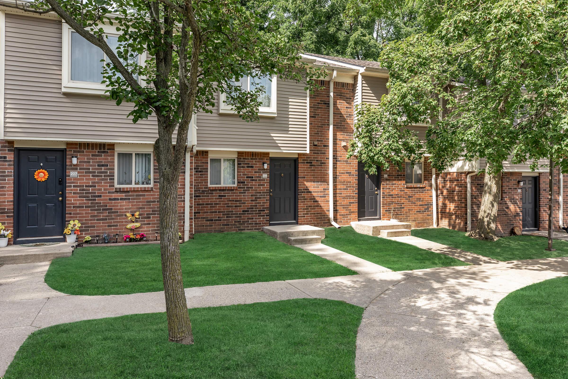 A row of townhouses with brick and beige siding. Each unit has a black door, and some have decorative fall wreaths. The pathway is lined with freshly cut grass and small flower beds. Trees provide shade, creating a welcoming neighborhood atmosphere. Sunlight casts gentle shadows on the walkways.