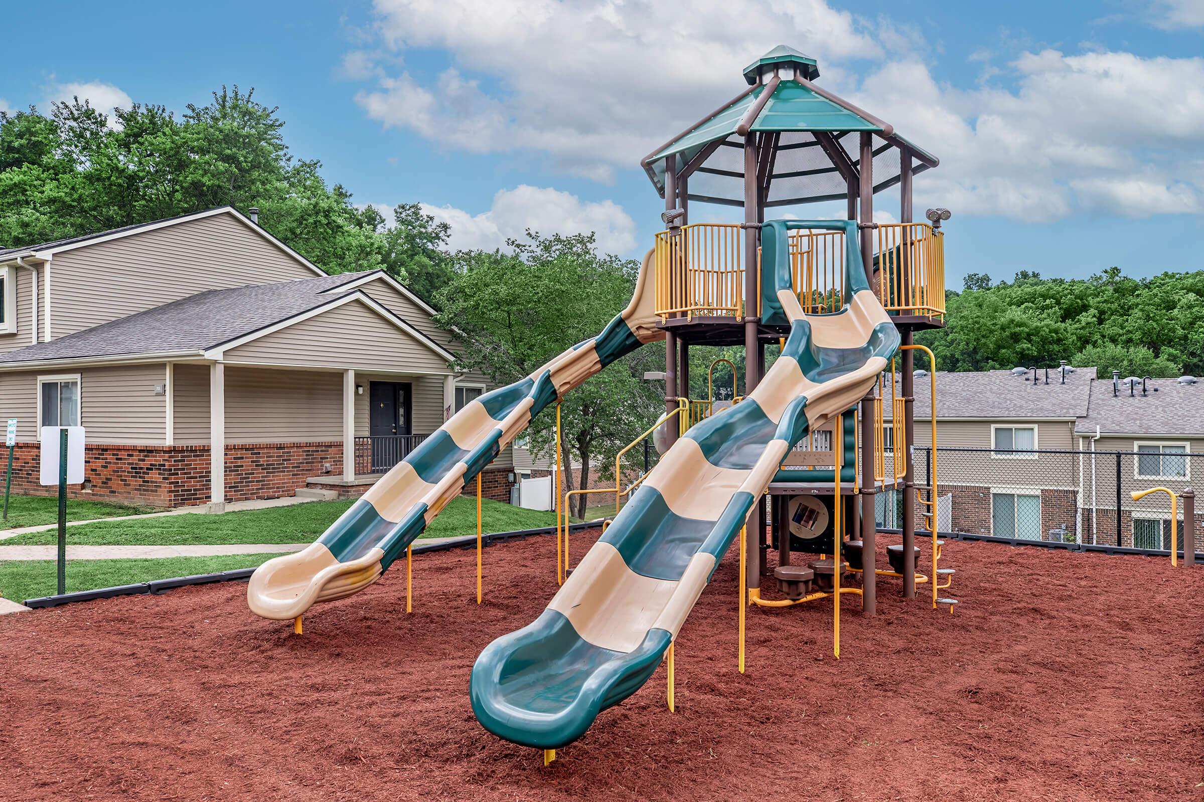 A playground featuring a multi-level structure with two green and beige slides, surrounded by red rubber mulch. Trees and residential buildings are visible in the background under a partly cloudy sky.