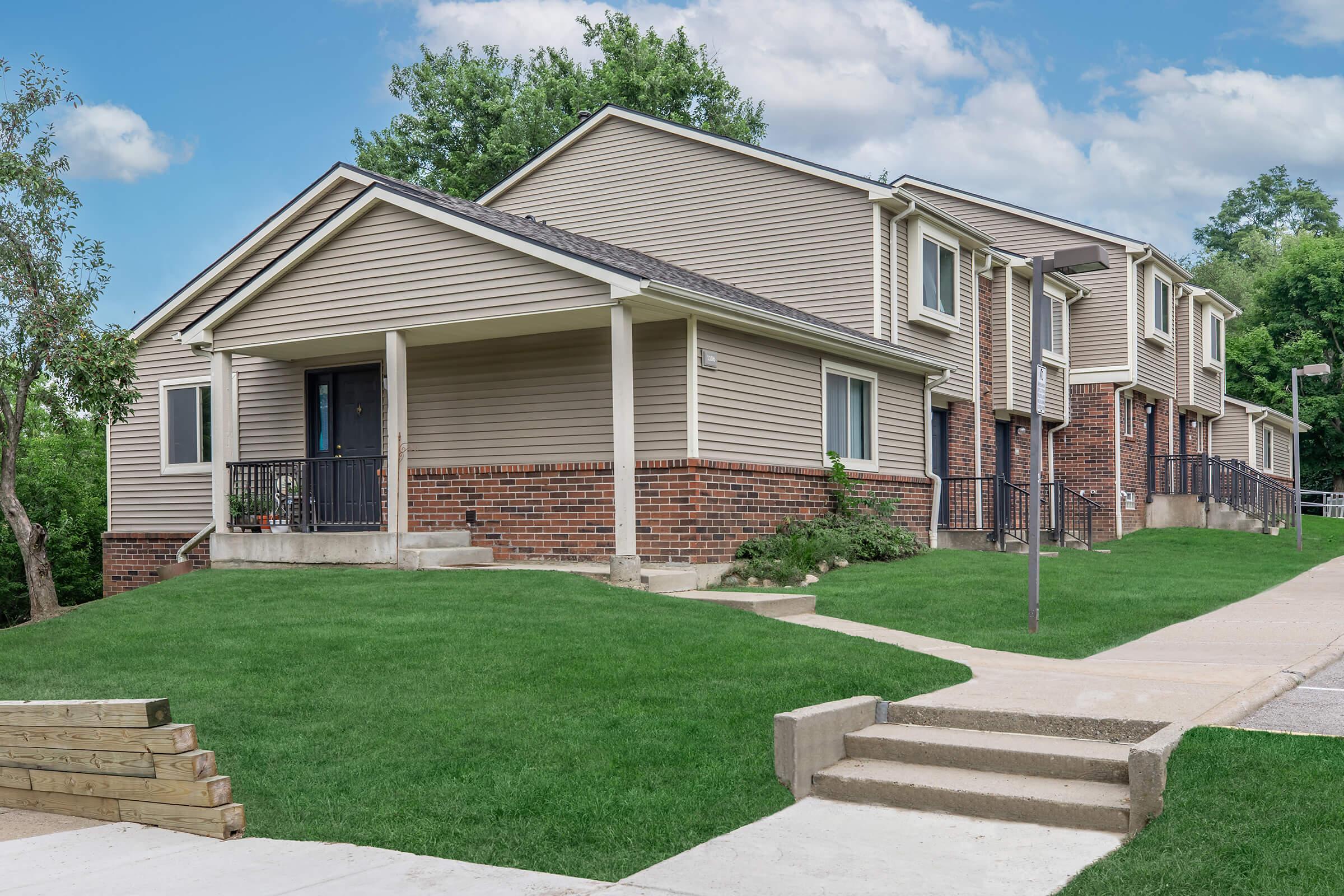 A modern two-story residential building with beige siding and brick accents, surrounded by green grass. Features include a walkway leading to a black door, multiple windows, and a sloped lawn. The environment is peaceful with trees in the background and a clear blue sky.