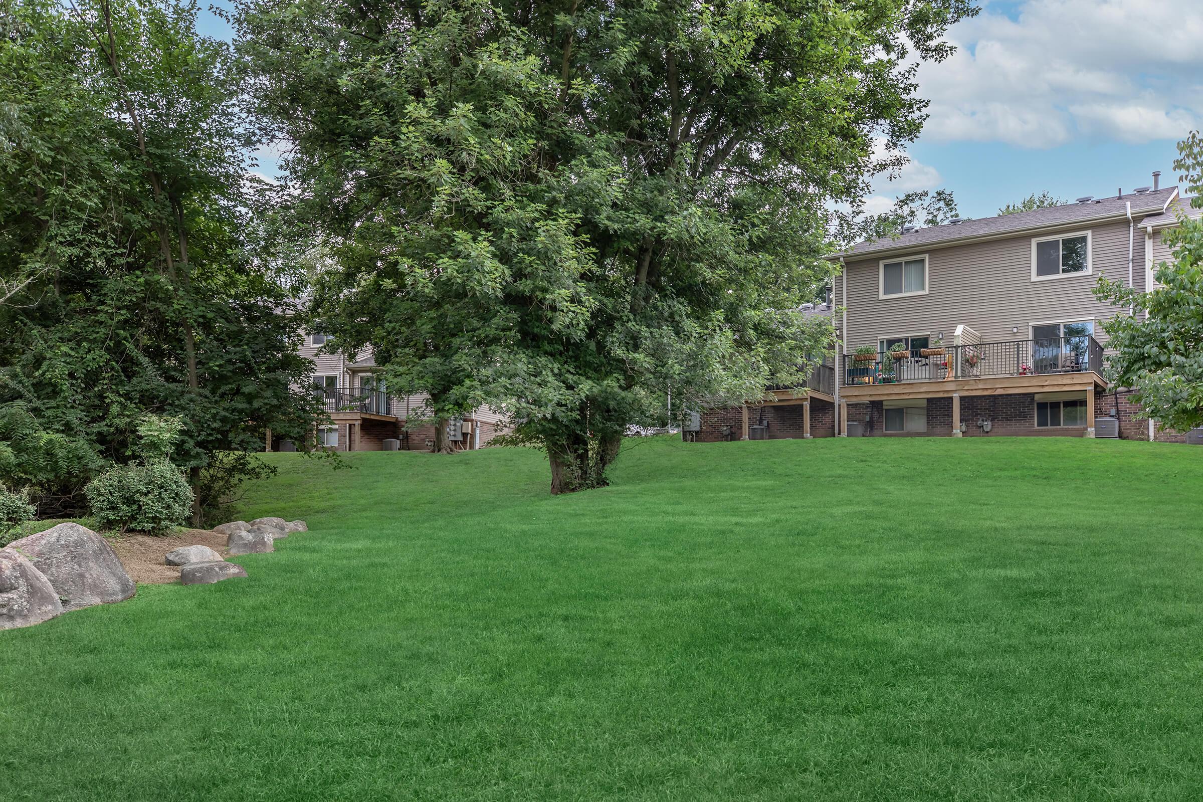Lush green lawn with a few large rocks scattered around, framed by trees. In the background, residential buildings are visible, featuring balconies and a landscaped area. The sky is partly cloudy, creating a serene outdoor setting.