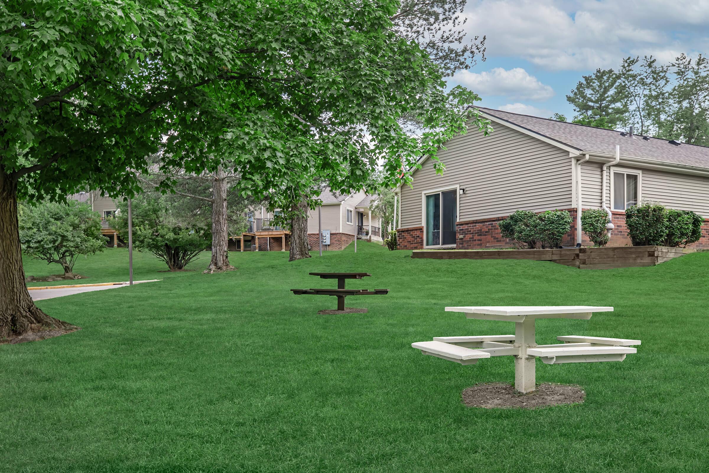 A green lawn with a white picnic table and a black circular table. In the background, there are two beige houses with brown accents, surrounded by trees and shrubs. The sky is partly cloudy, creating a tranquil outdoor setting.